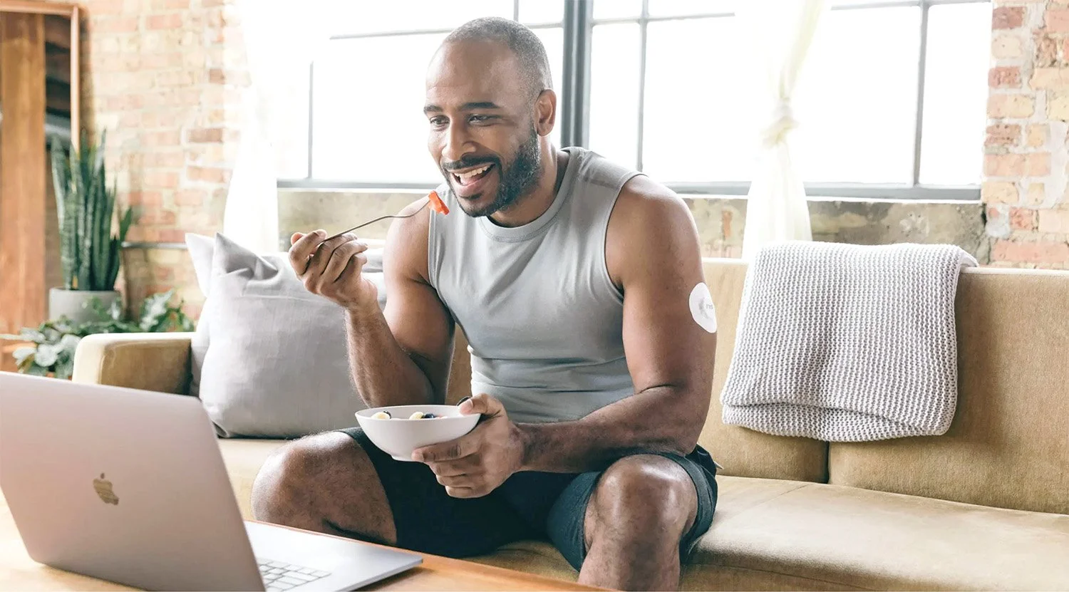 Fit man sits on couch, eating fruit as he takes part in a conversation via his laptop.