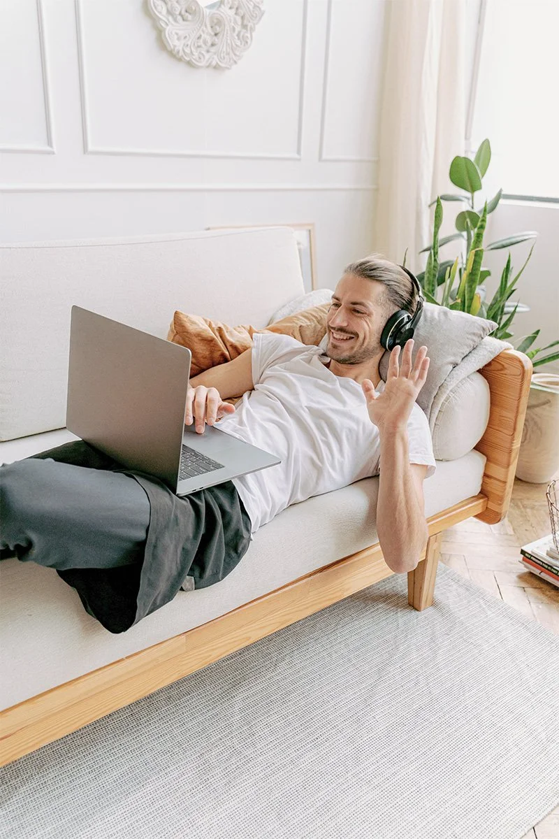 Man laying on couch, wearing headphones, waving at people on his laptop