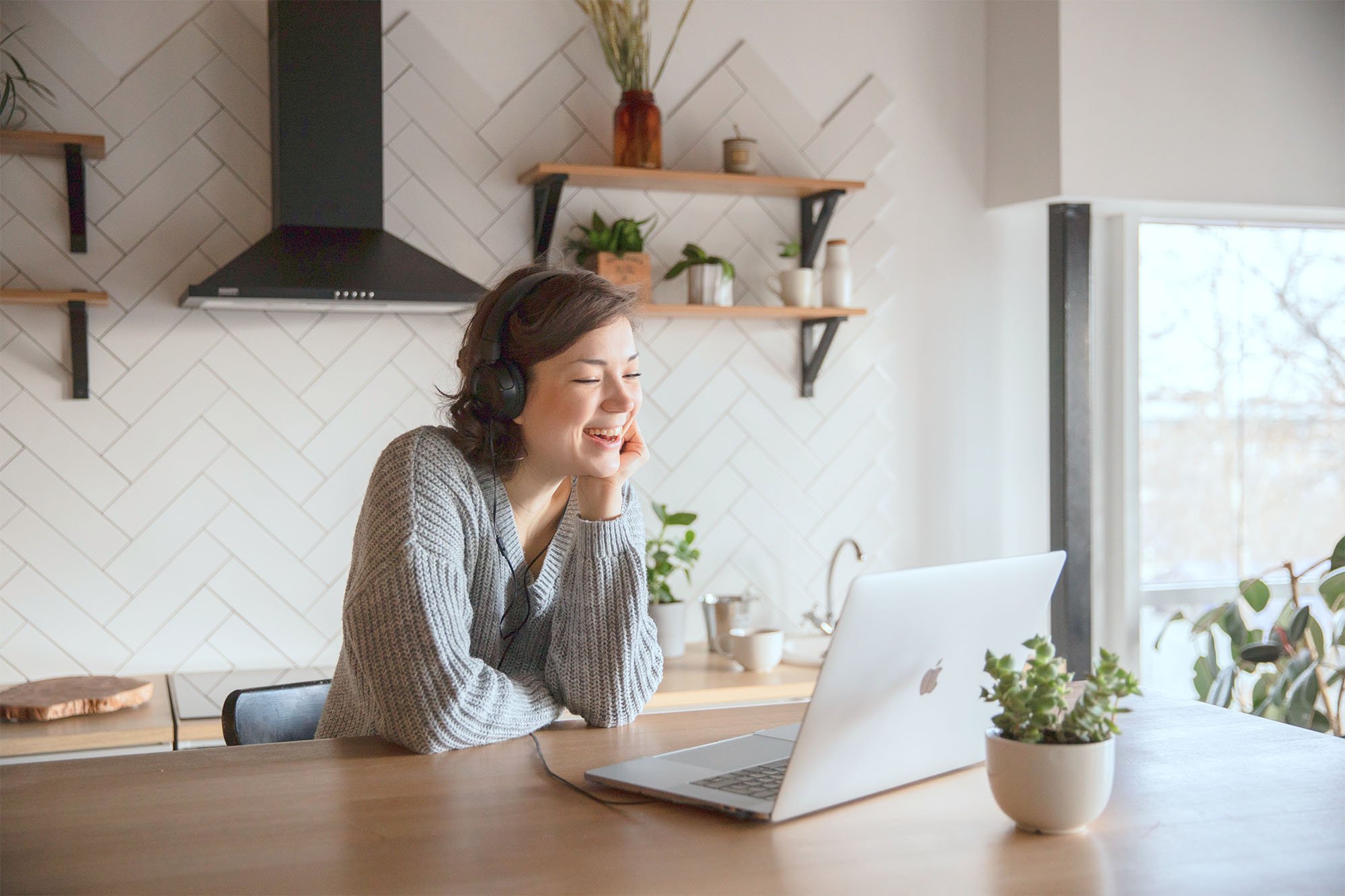 Young woman sits at kitchen counter, smiling and talking to someone on her laptop.