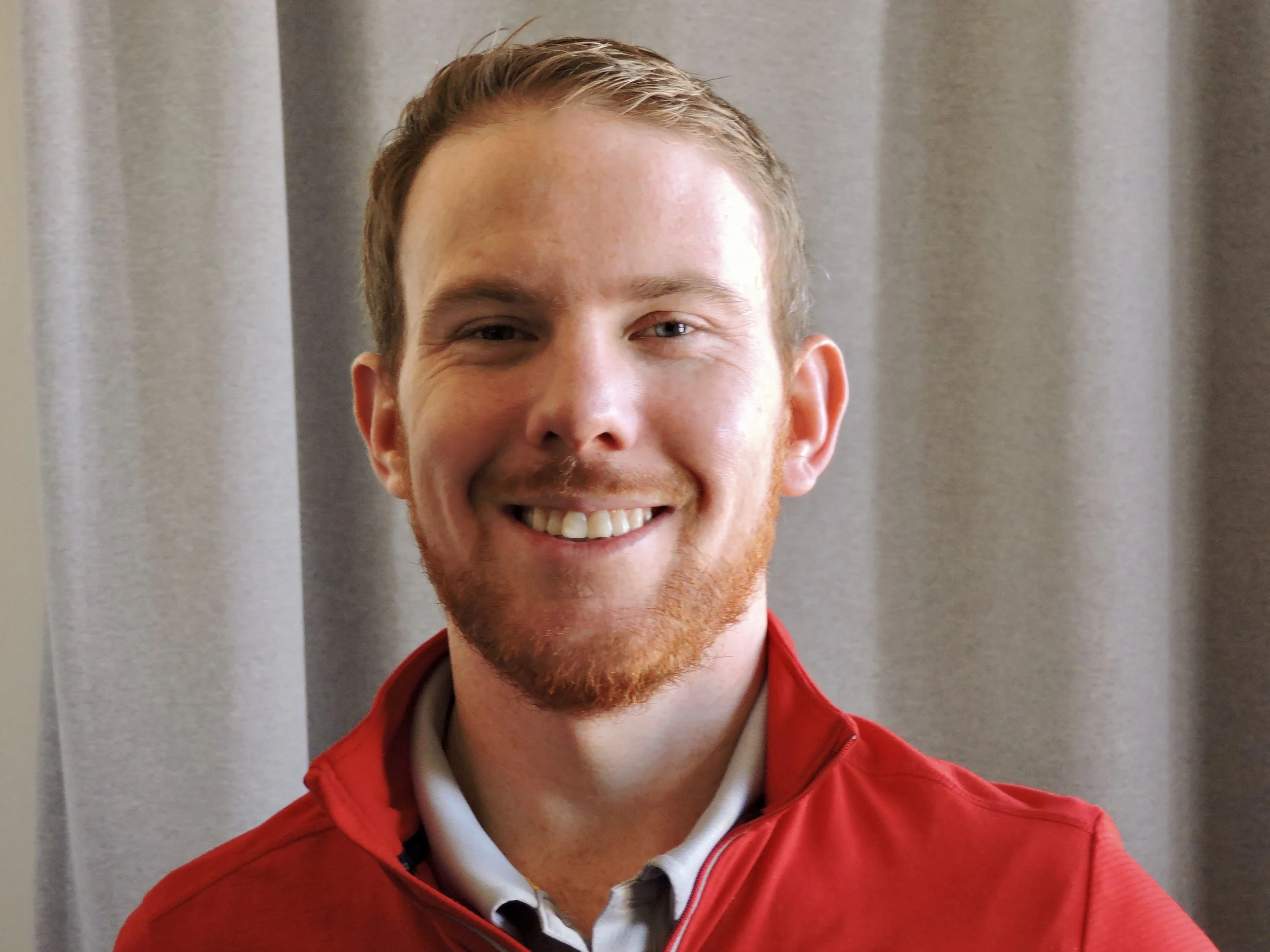 A young man with short red hair, a beard, and a big smile, wearing a red jacket and a white collared shirt, standing in front of a gray curtain.