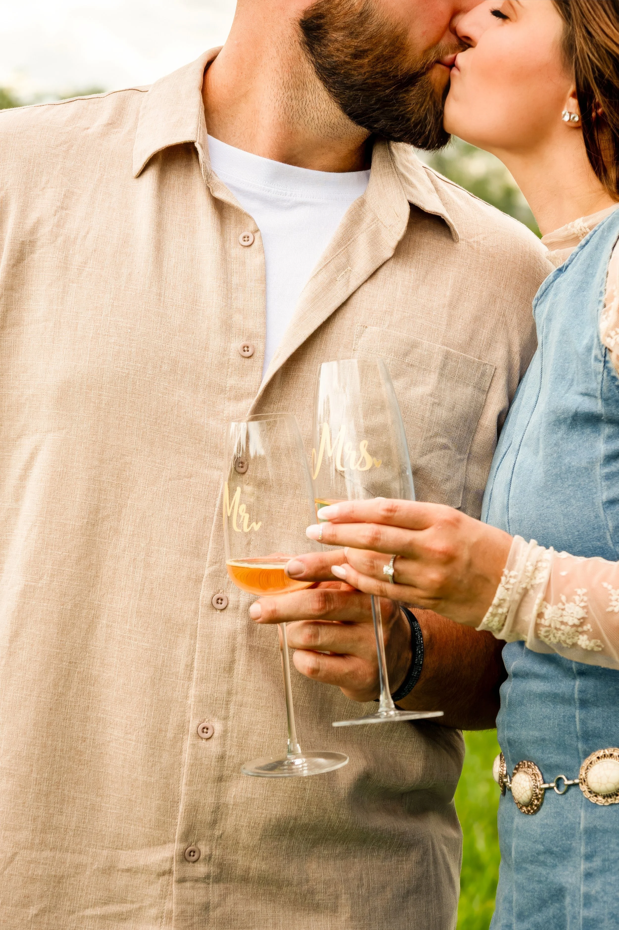 A close-up of a couple kissing outdoors during daytime, holding wine glasses with 'Mrs.' written on them. The man is wearing a beige button-up shirt with a white T-shirt underneath, and the woman is wearing a light blue dress with lace and pearl deta