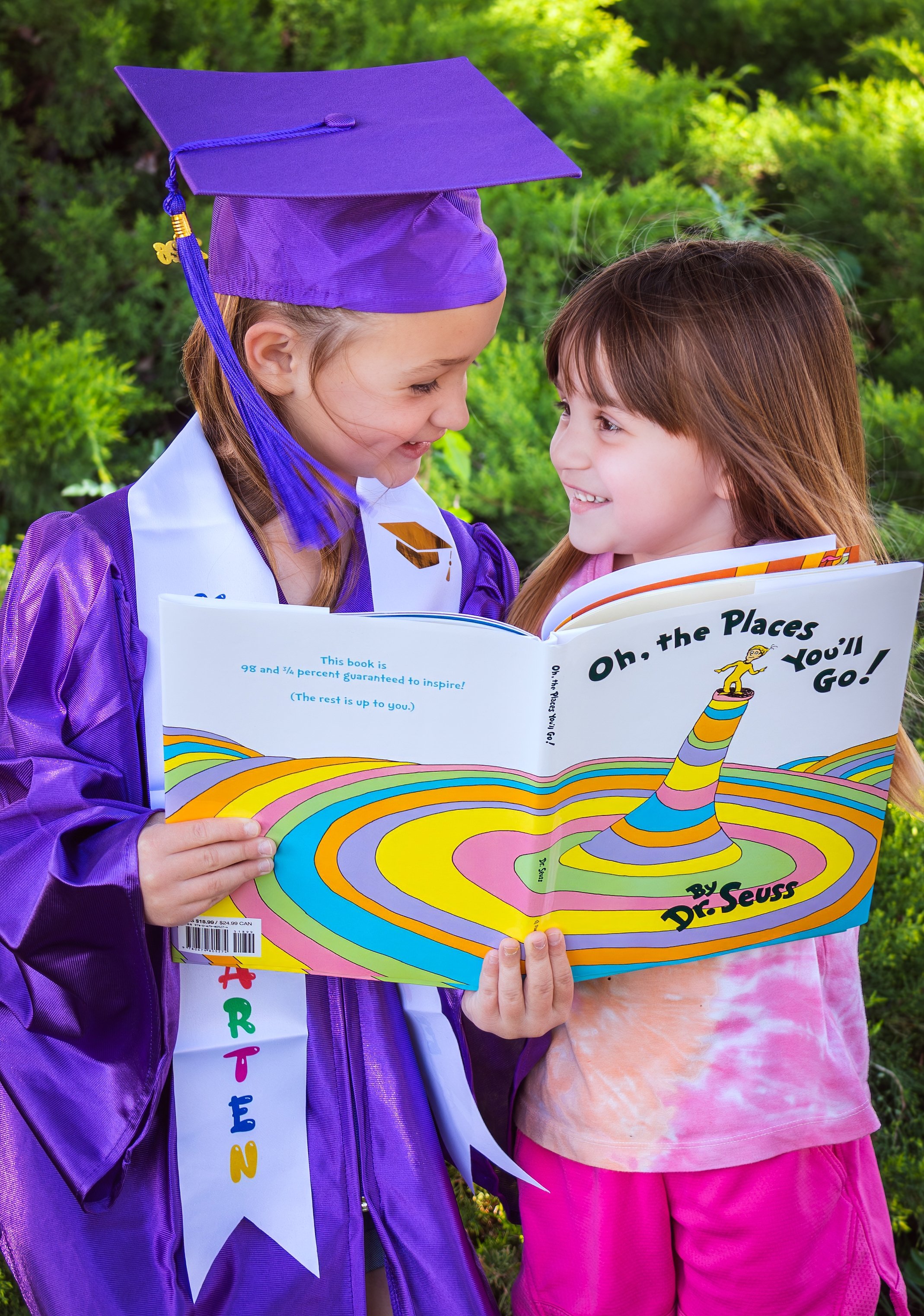 Two young girls in purple graduation gown and pink outfit, smiling and reading a colorful "Oh, the Places You'll Go!" Dr. Seuss book outdoors.
