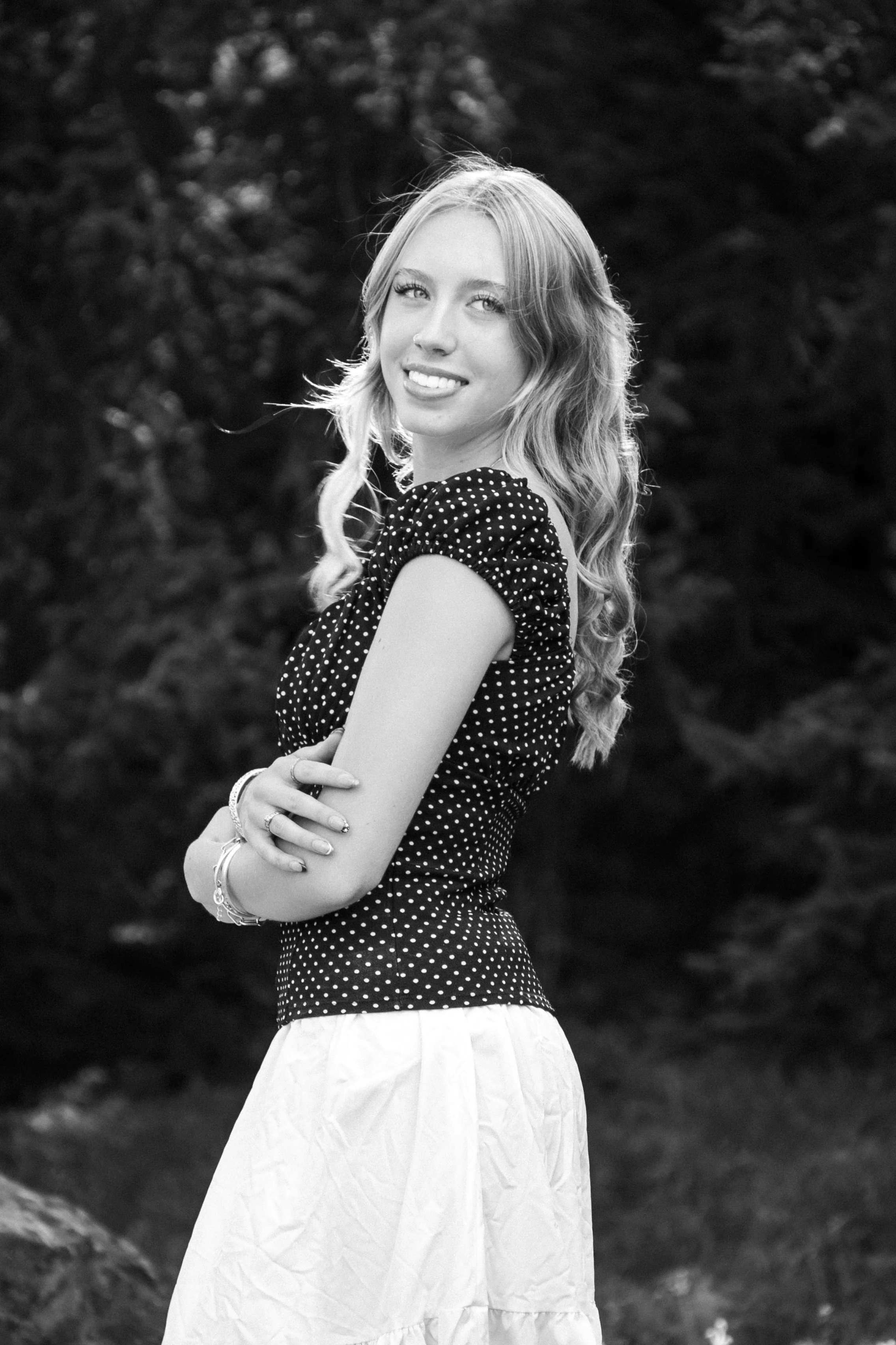 A young woman with wavy hair and a bright smile, wearing a polka dot top and a light-colored skirt, standing outdoors in front of a blurred natural background.