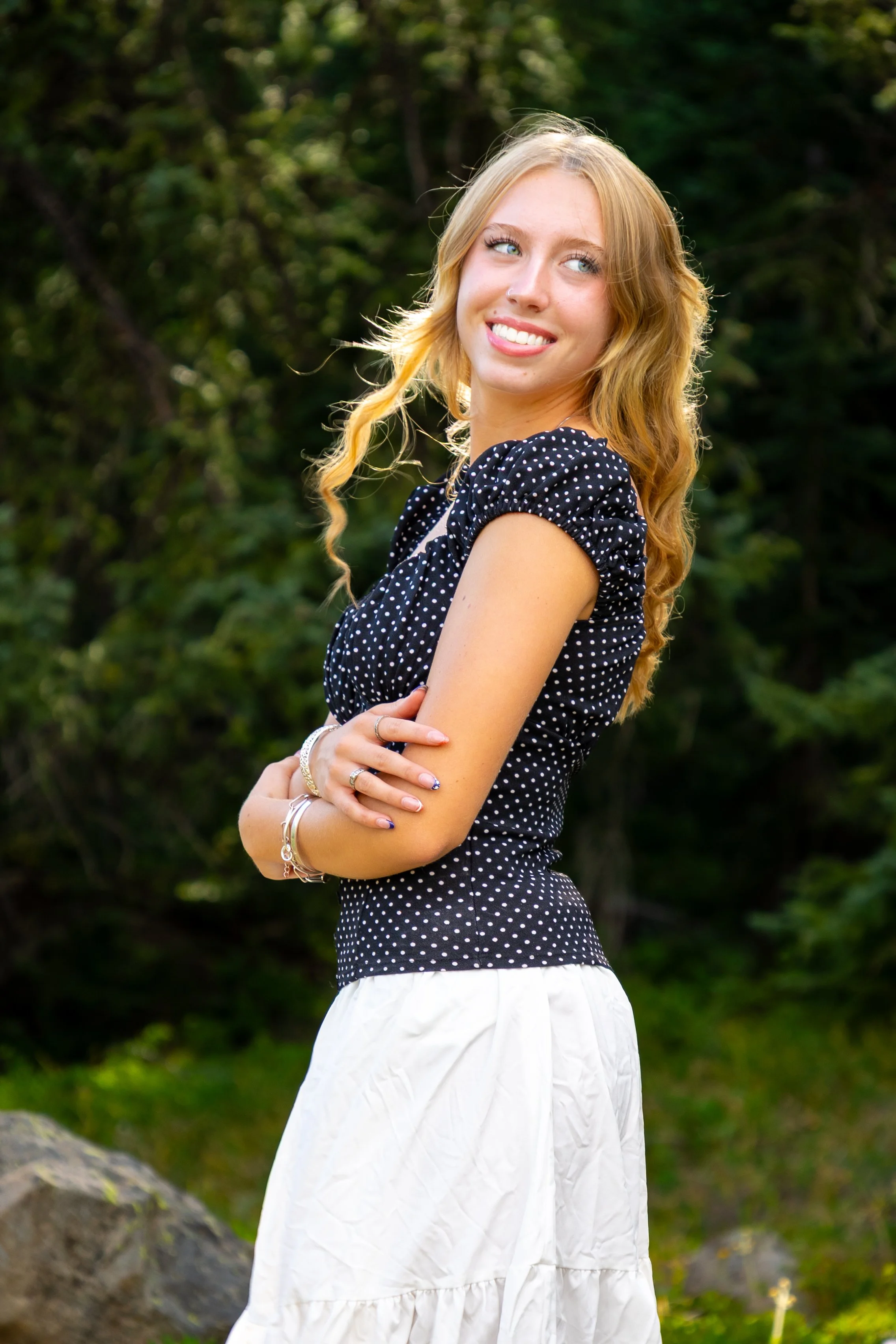 A young woman with long wavy blonde hair, blue eyes, and a nose piercing, smiling and looking to the side, standing outdoors with green trees in the background. She is wearing a black polka dot short-sleeve top and a white skirt, accessorized with je