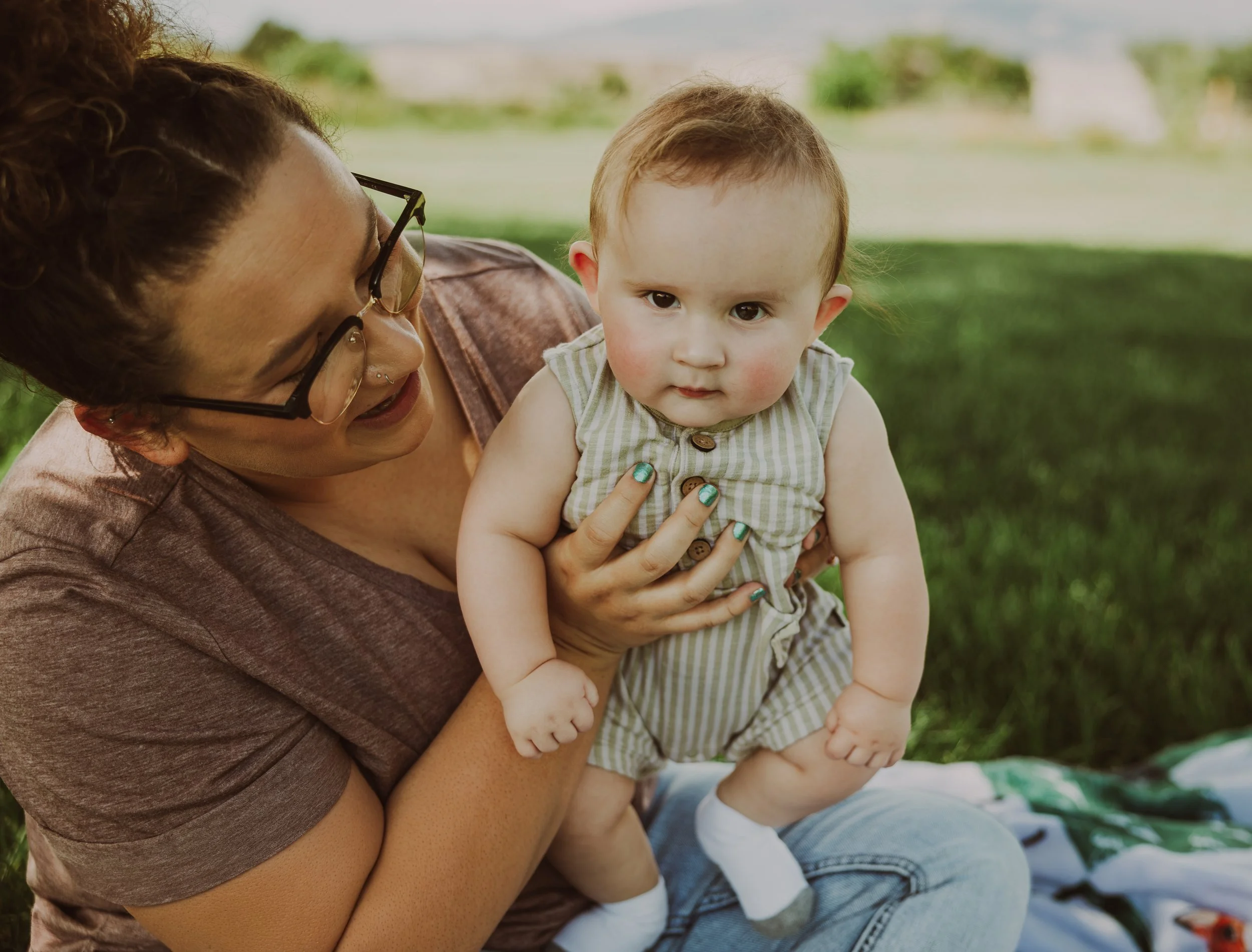 A woman holding a young boy outdoors on a grassy area, with blurred green trees and sky in the background.