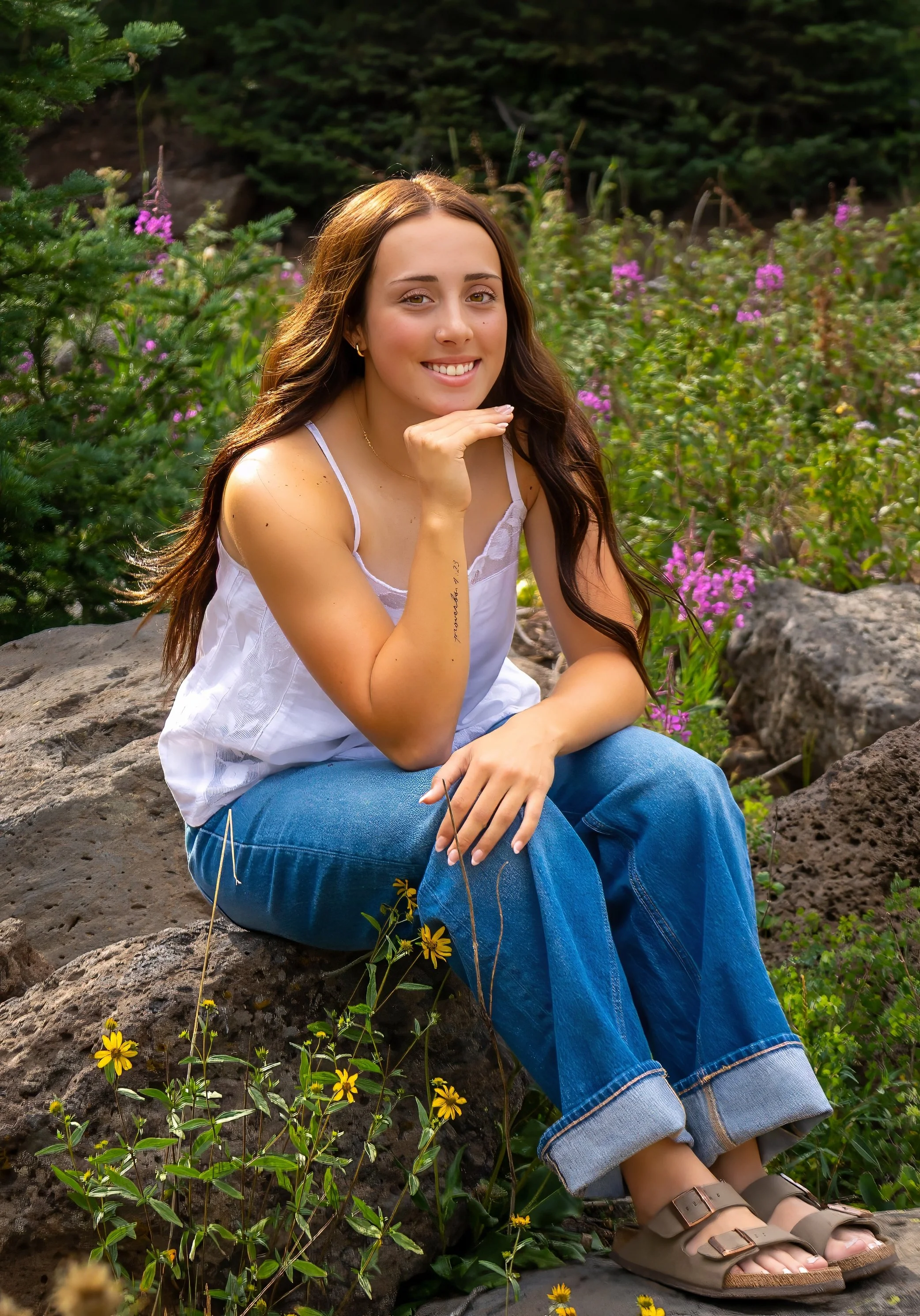 Young woman with long brown hair smiling, sitting on rocks among greenery and yellow flowers, wearing a white top, blue jeans, and sandals.