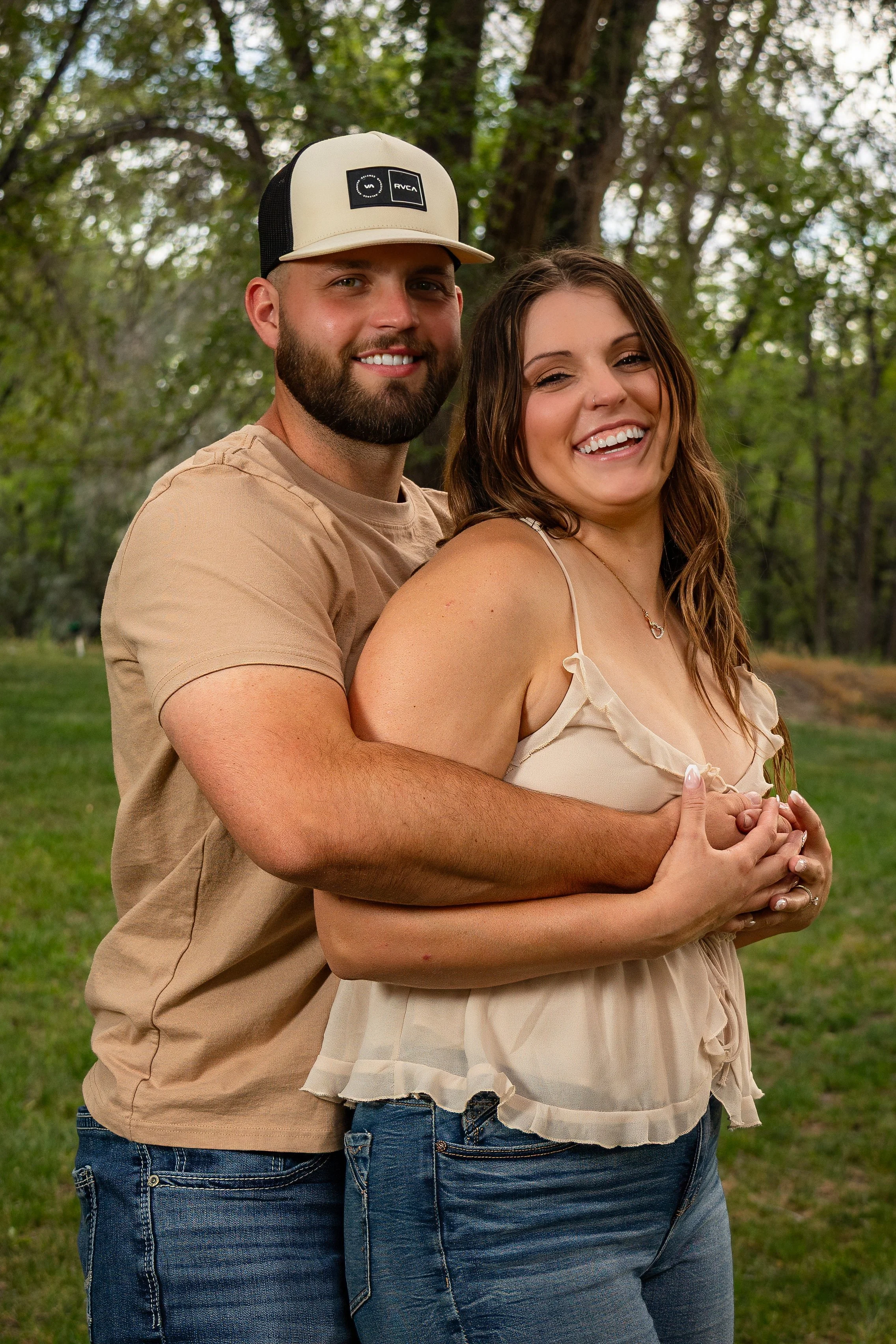 A smiling man and woman embracing outdoors in a park with trees in the background.