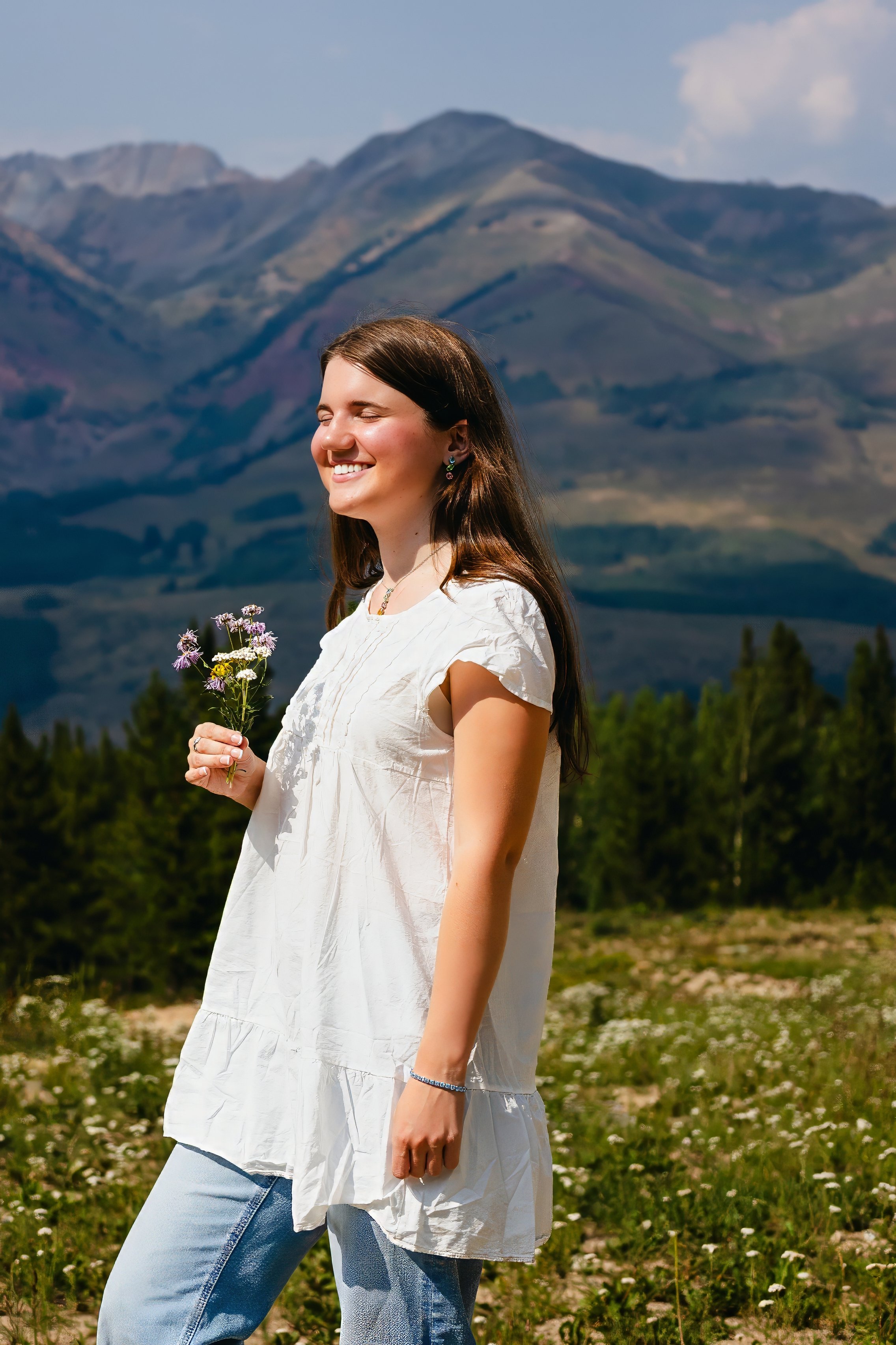 A woman smiling with closed eyes, holding a small bunch of wildflowers, standing outdoors in a field of small white flowers with mountains in the background on a clear day.