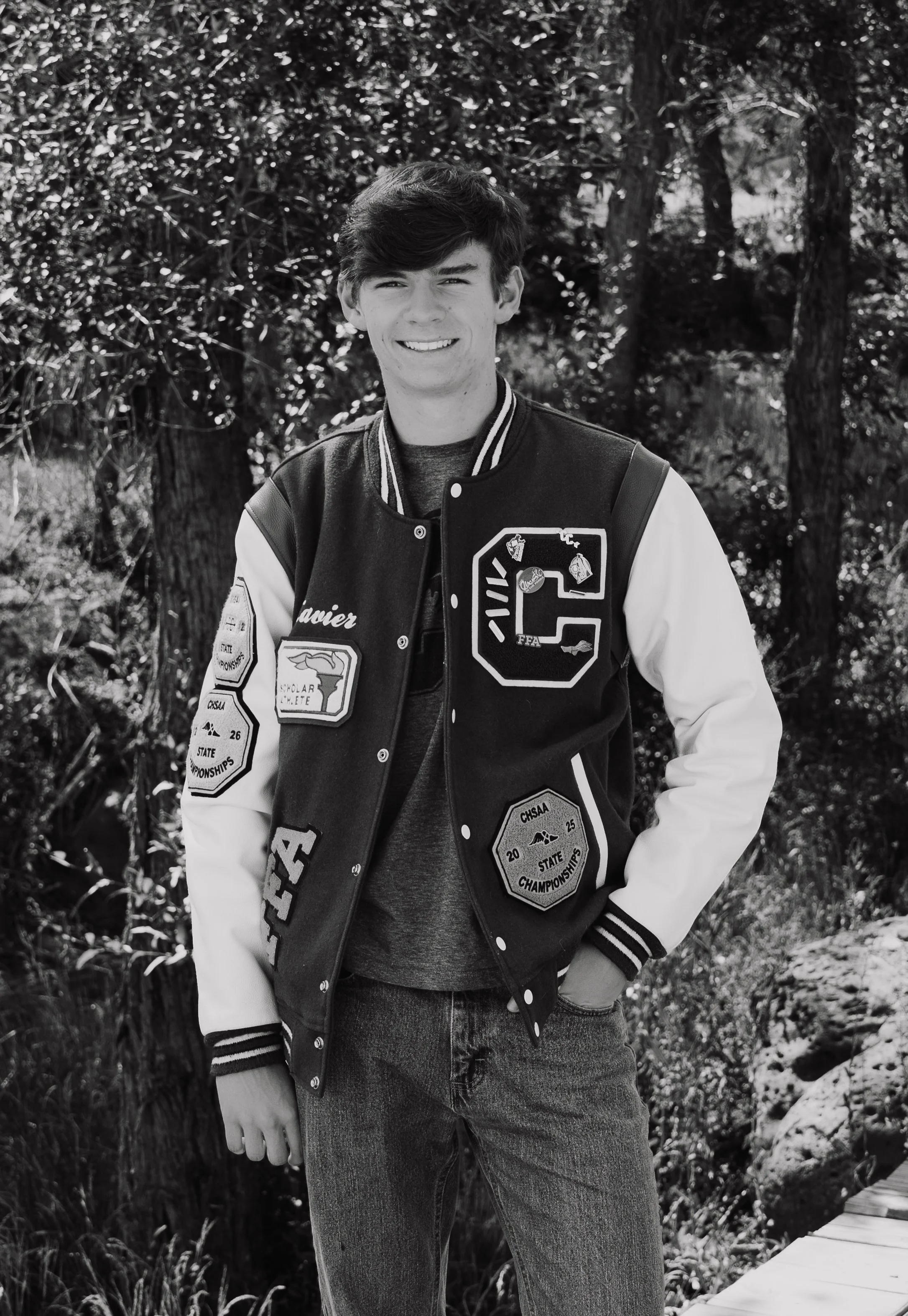 A young man with dark hair smiling outdoors in front of trees and bushes, wearing a varsity jacket with patches and jeans.
