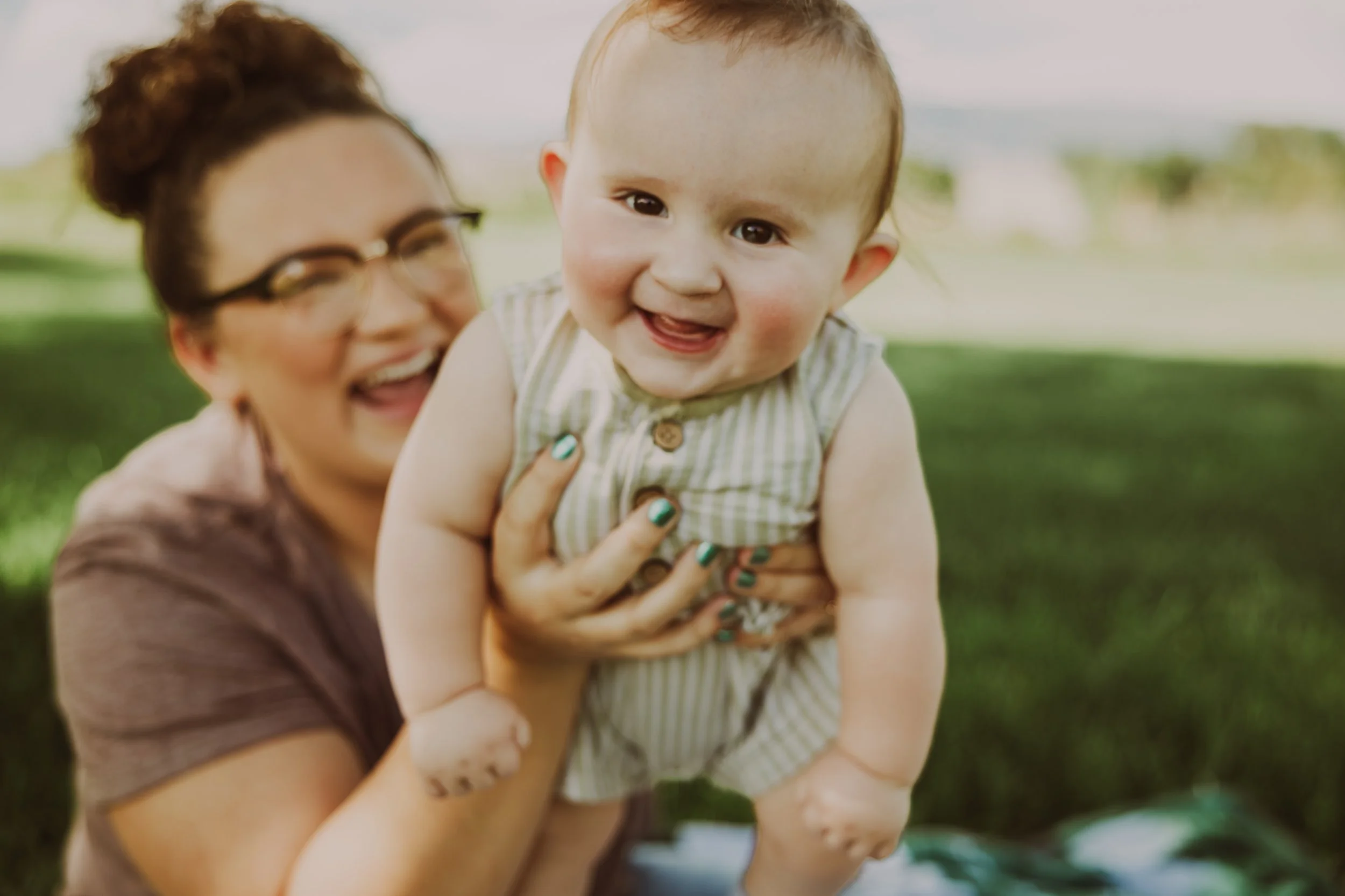 A woman holding a happy smiling toddler outdoors with green grass and trees in the background.