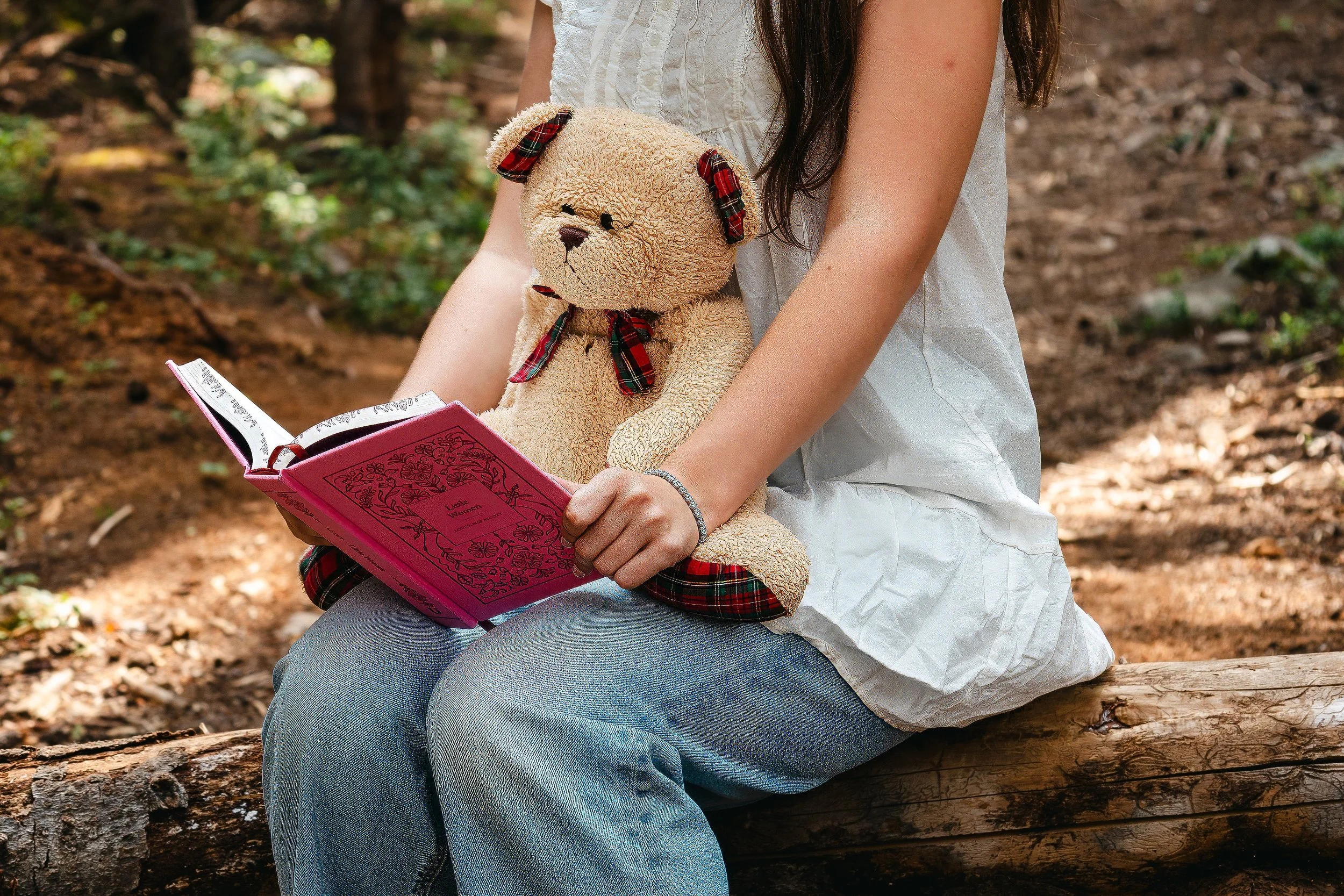 A person sitting outdoors on a log, holding a teddy bear with a plaid ribbon and ears, and reading a pink book titled "Love Wins".
