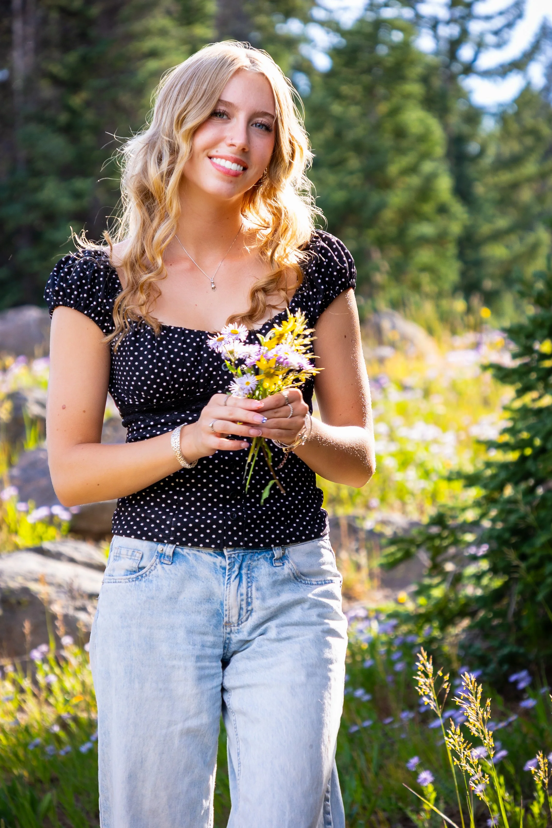 A woman with long blonde hair, wearing a black polka dot top and light blue jeans, holding a small bouquet of flowers outdoors in a sunny, green natural setting.