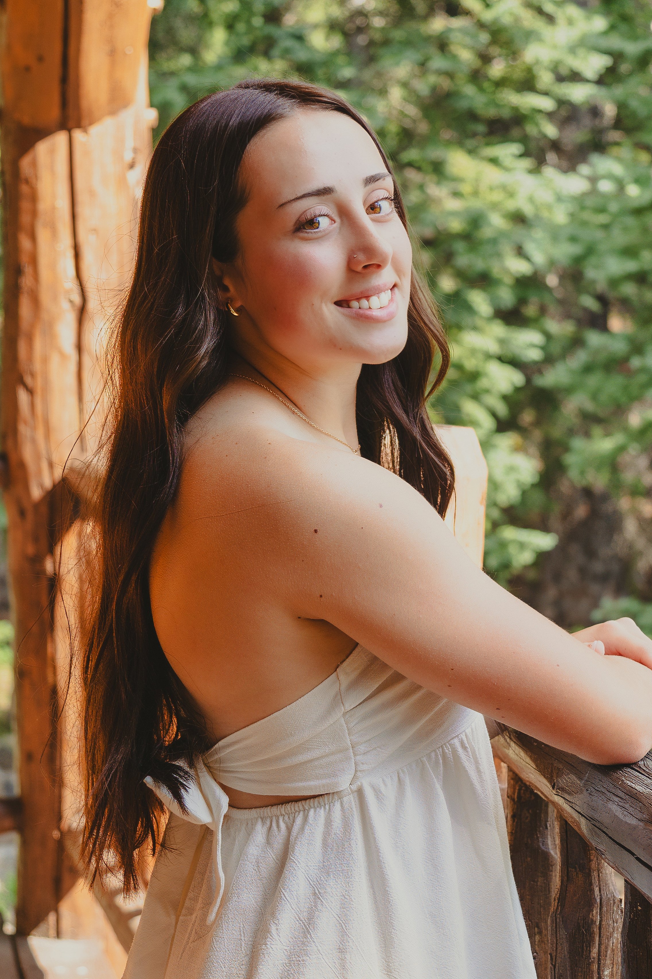 A young woman with long dark hair, wearing a beige strapless dress and a small nose piercing, standing outdoors on a wooden structure with green trees in the background. She is smiling and looking at the camera.