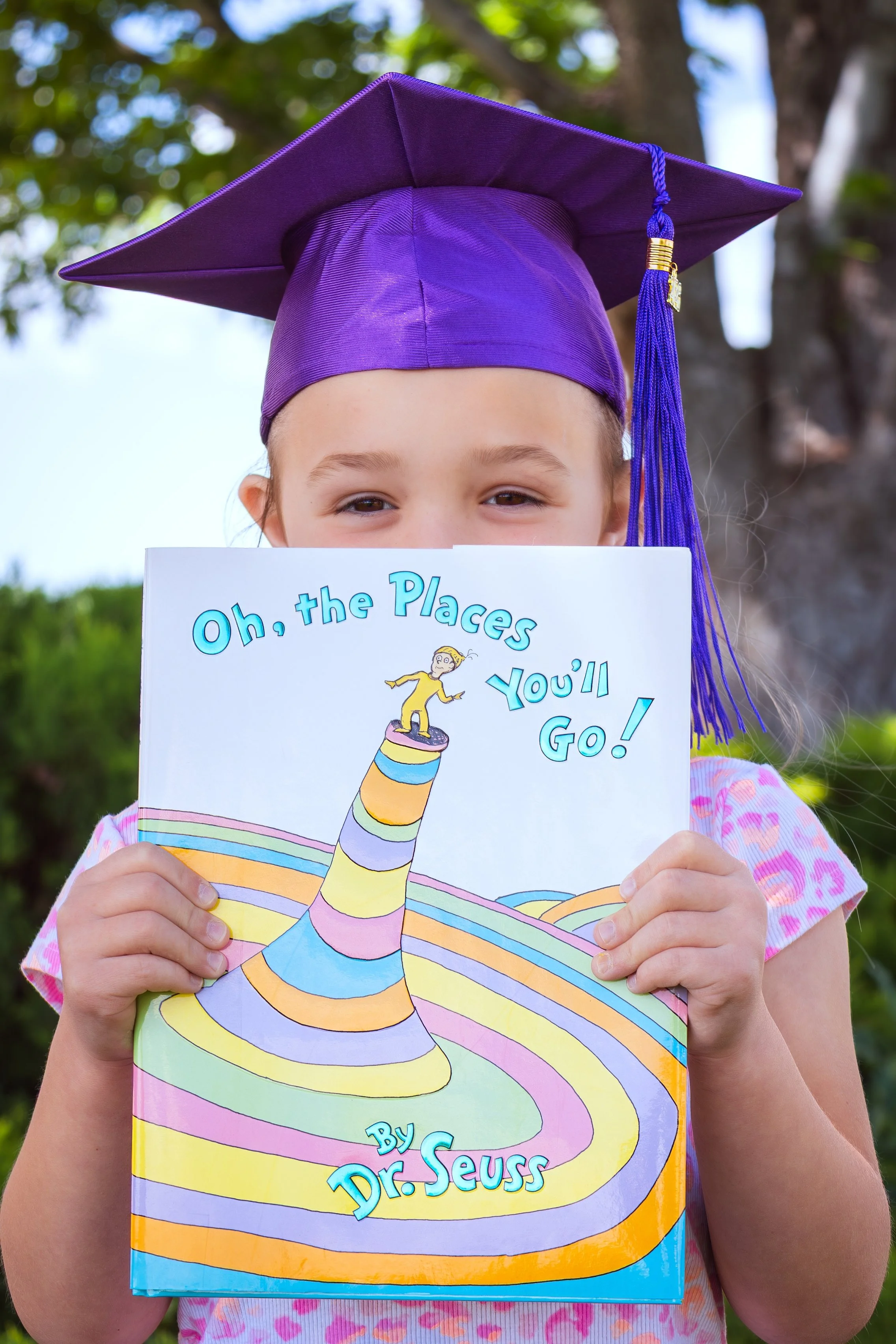 A young girl wearing a purple graduation cap and holding a colorful Dr. Seuss book called "Oh, the Places You'll Go!" outdoors with a blurred background of trees and sky.