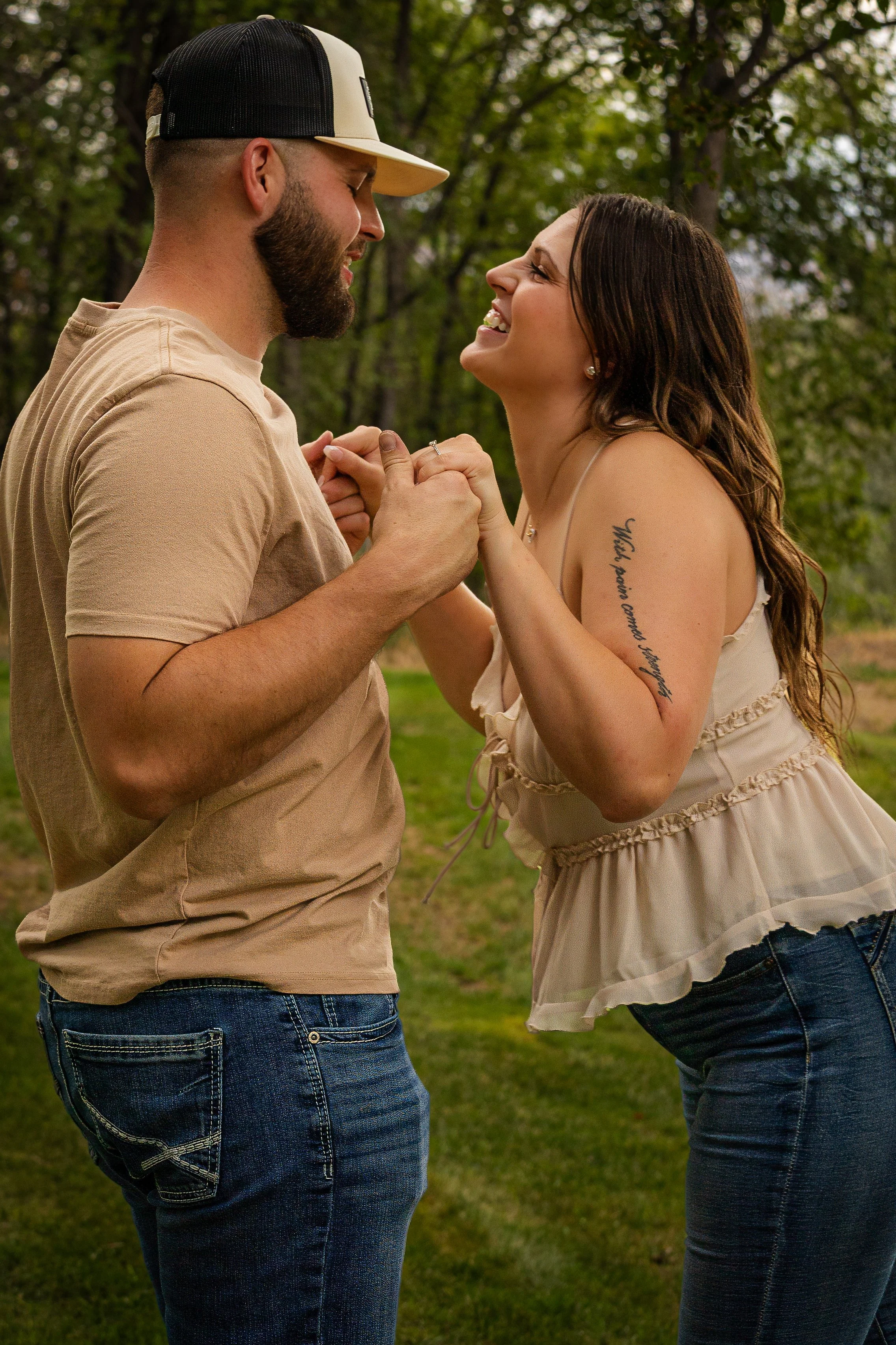A couple holding hands and smiling at each other outdoors in a park, surrounded by green trees.
