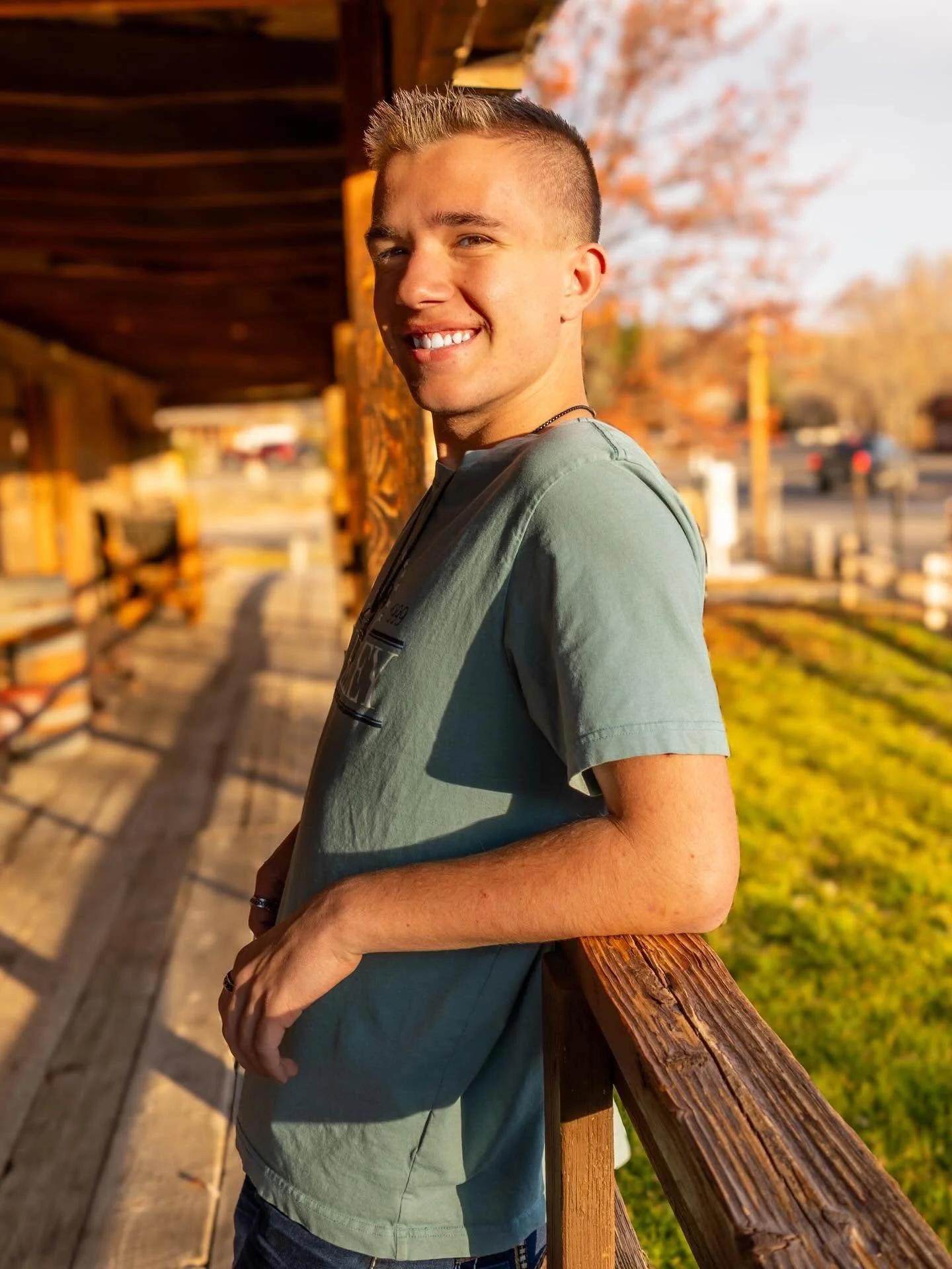 A young man with short hair standing outside on a wooden porch, smiling at the camera, with trees and a parking lot in the background during sunset.