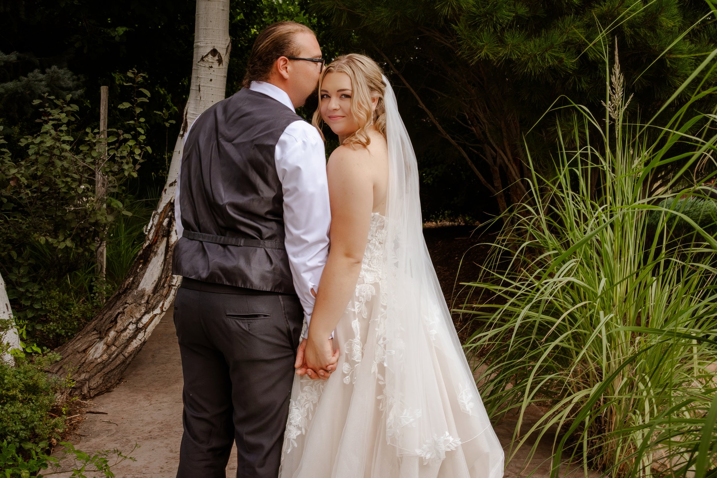 A bride and groom holding hands in an outdoor garden setting, surrounded by trees and green plants, with the bride smiling and looking at the camera.