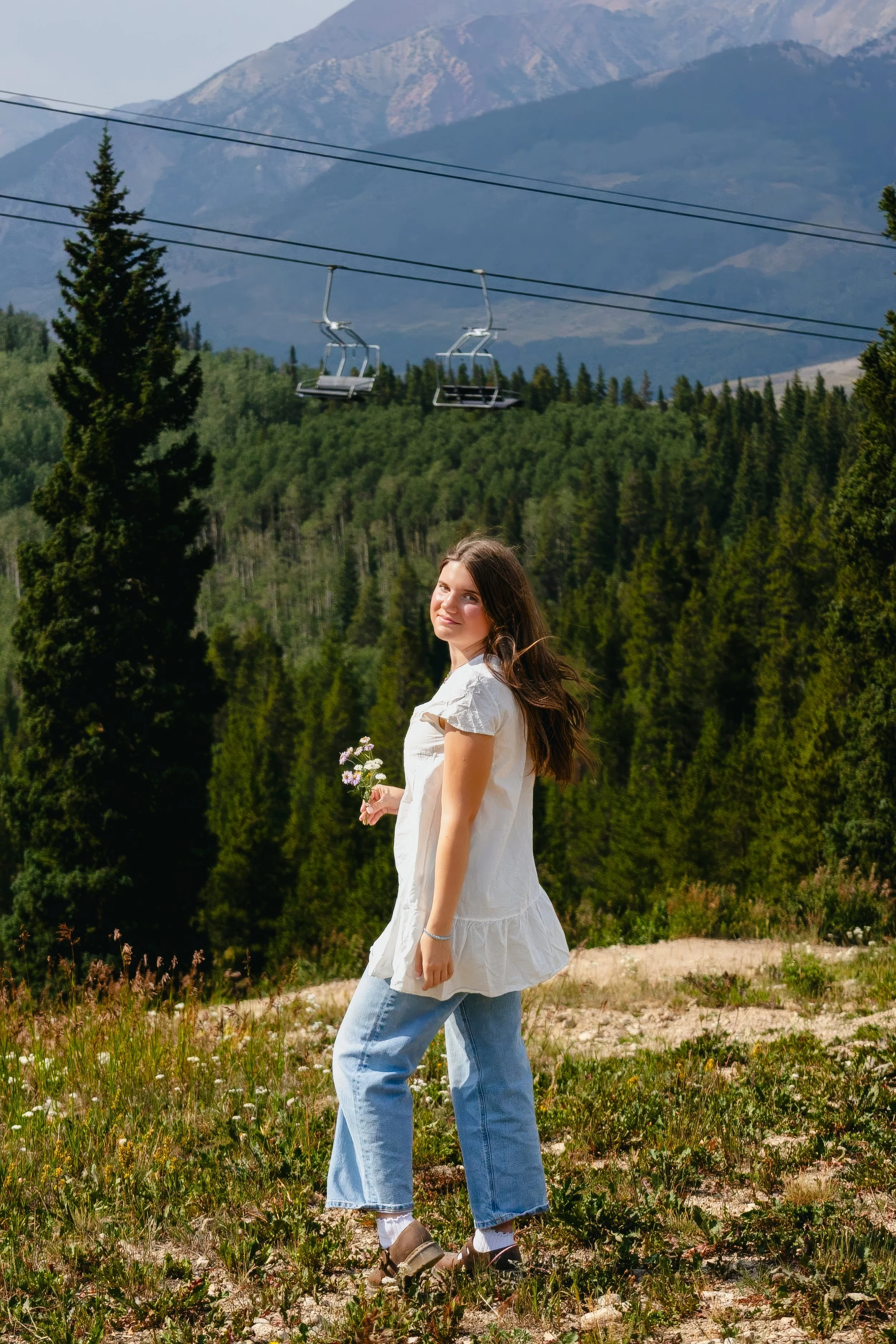 A young woman with long brown hair wearing a white dress and jeans, holding a small bouquet of flowers, standing in a field with mountains and trees in the background under a clear sky.