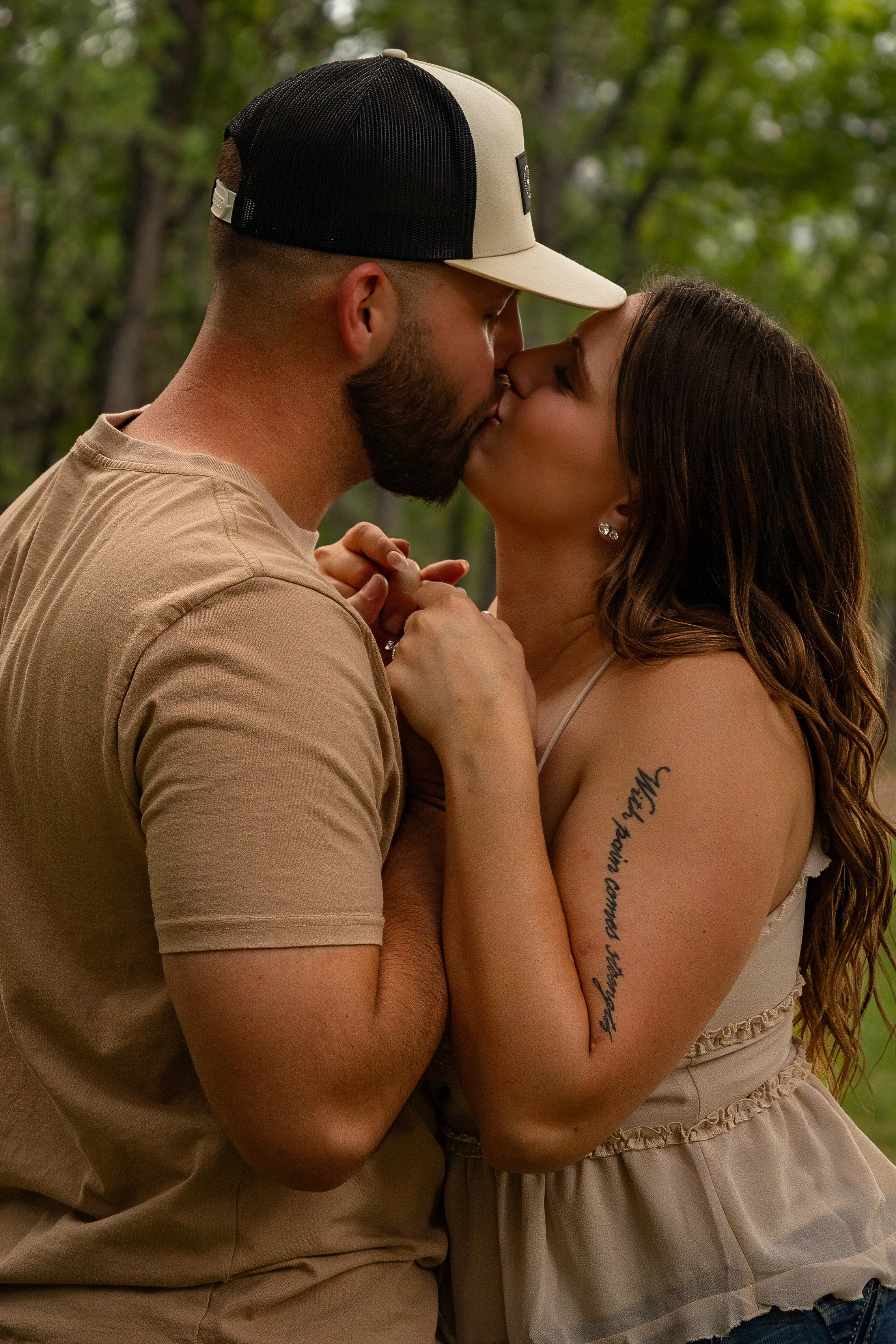 A man and woman sharing a kiss outdoors, with trees in the background. The man is wearing a beige t-shirt and a black and beige baseball cap, while the woman has long brown hair and is wearing a beige dress with ruffles. She has a tattoo on her left 