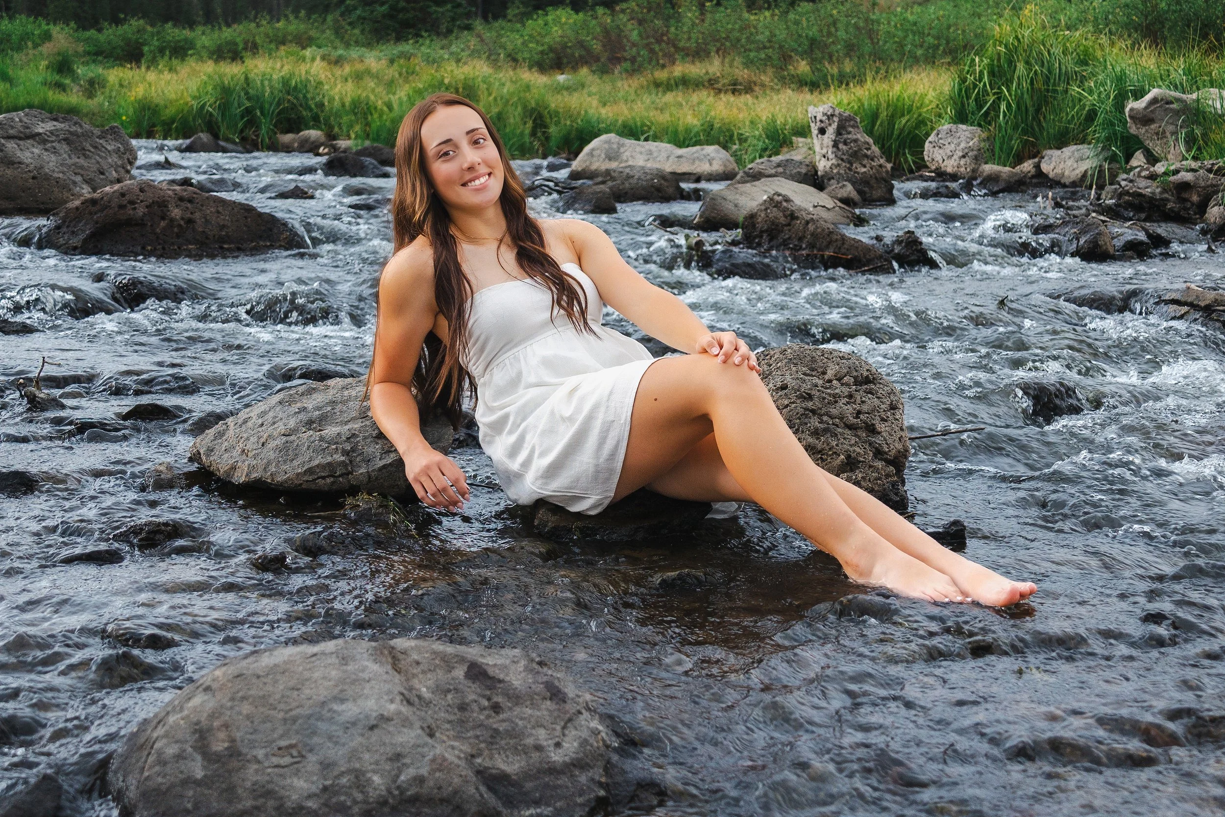 A young woman with long brown hair and a white dress sitting on rocks in a river, smiling at the camera, surrounded by greenery.