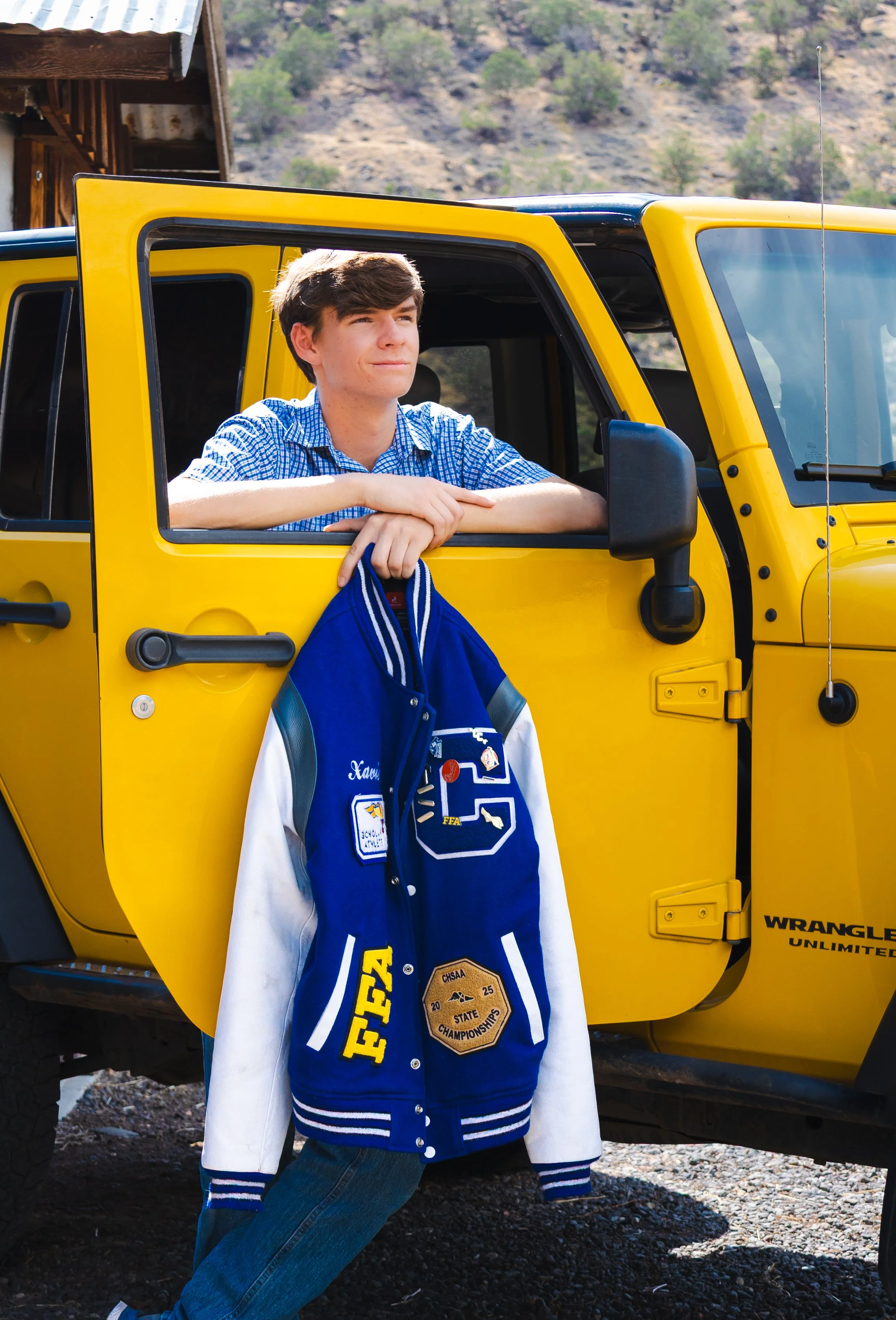 Young man with brown hair leaning out of a yellow Jeep Wrangler, wearing a blue varsity jacket with patches, jeans, and a blue shirt underneath. The Jeep has a black side mirror and the background shows a hillside with trees.