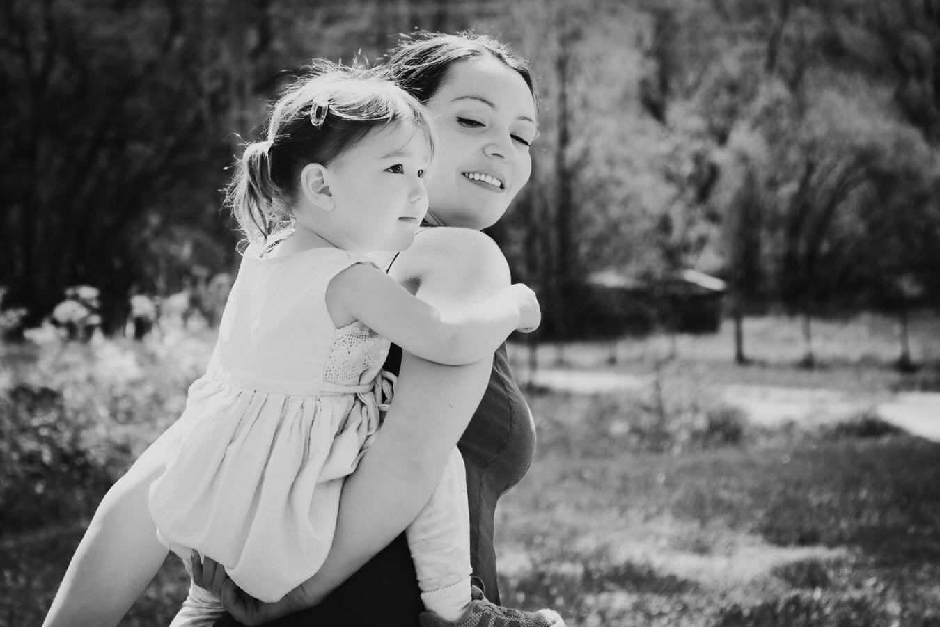 A woman carrying a young girl on her back outdoors in a park, both smiling and enjoying the moment.