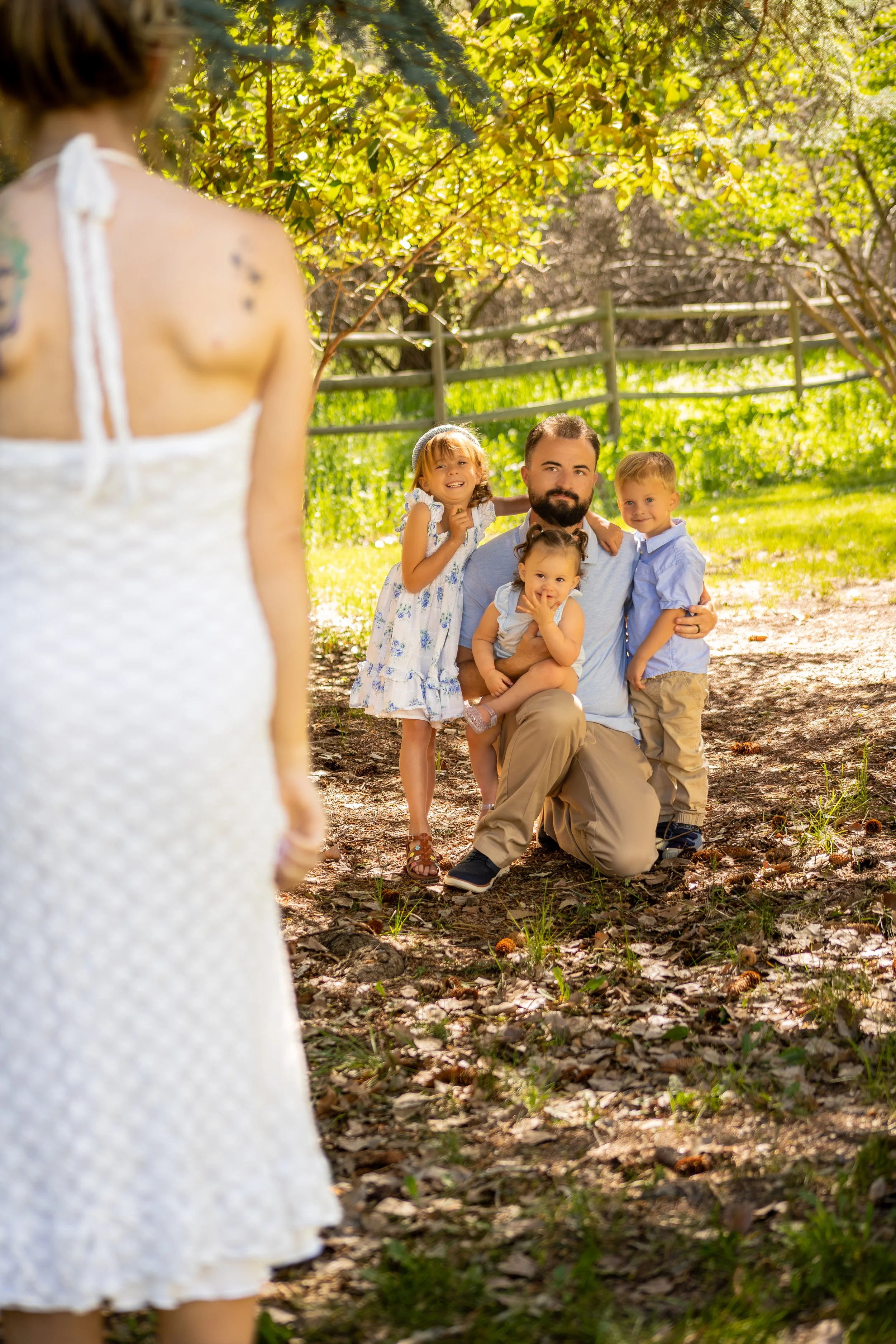A woman in a white dress with her back to the camera looking at a man and three children posing for a photo outdoors among trees and grass during the daytime.