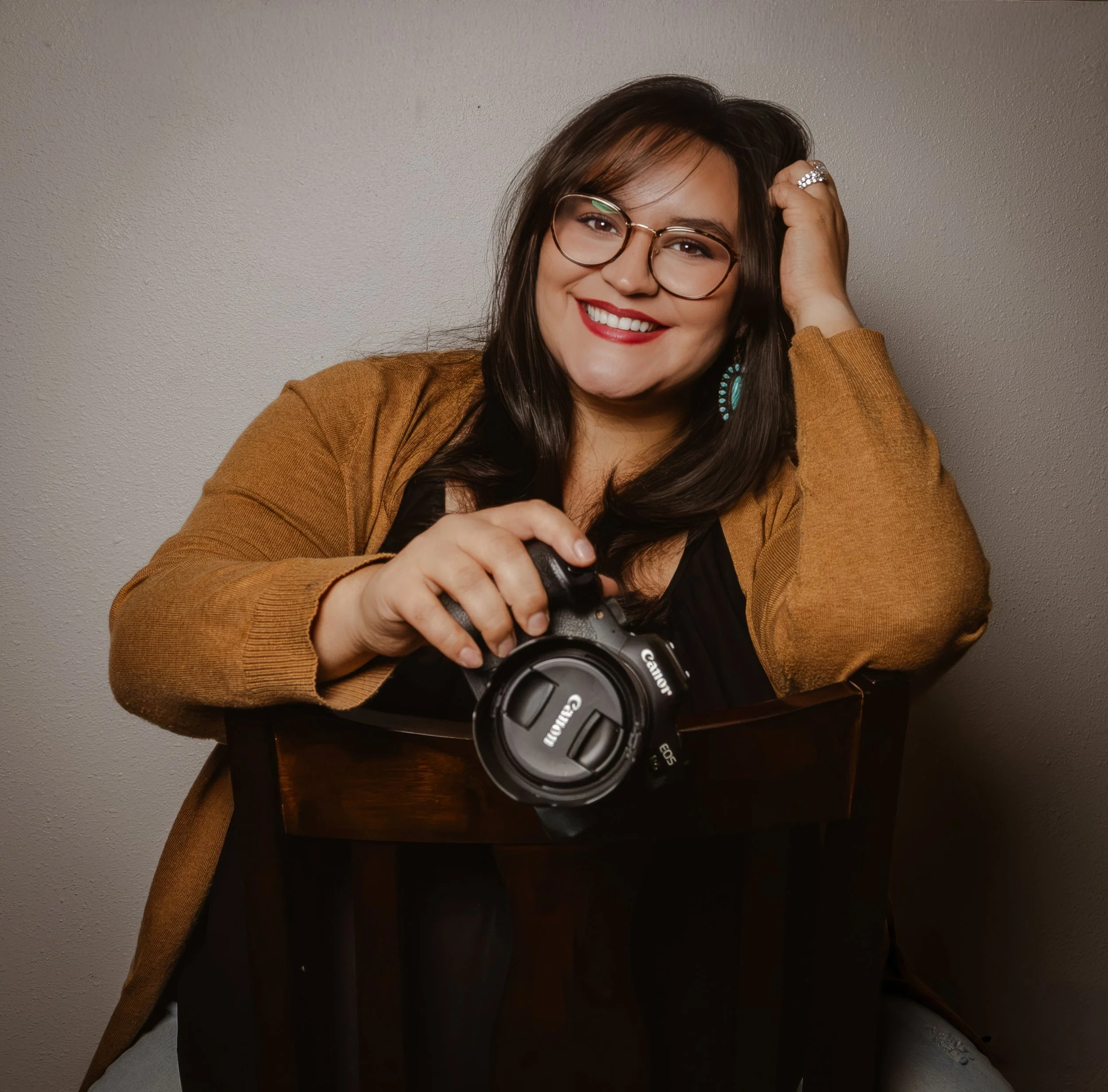 A woman with long dark hair wearing glasses, red lipstick, large earrings, a tan jacket, and a black top, smiling while holding a camera in front of a plain gray wall.