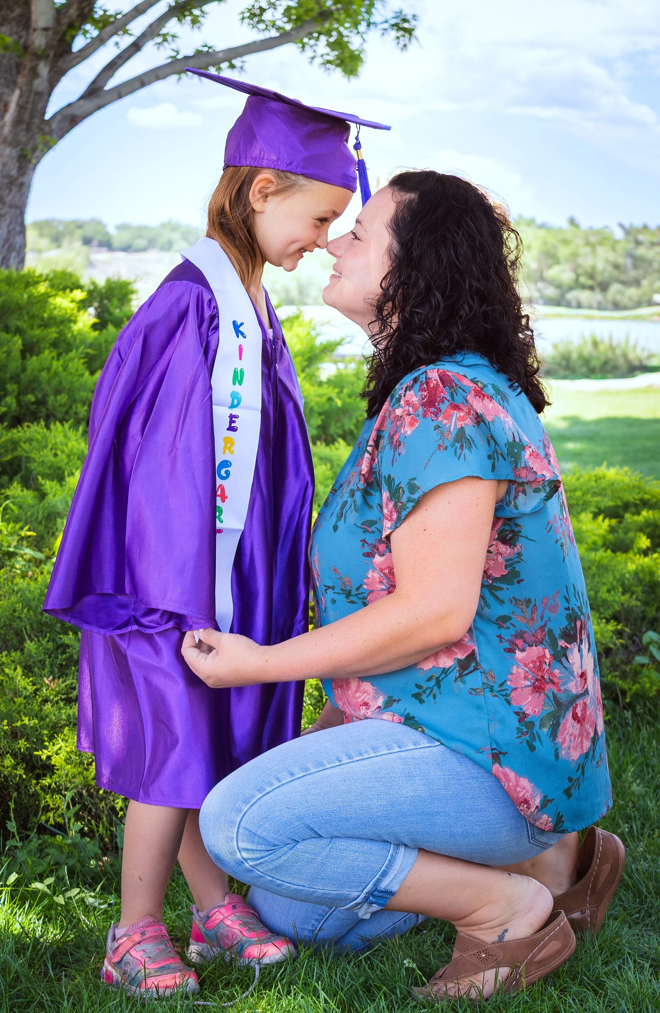 A young girl in a purple graduation cap and gown with a personalized sash and pink sneakers kneels to face her mother, who is kneeling and holding her hands, outside on a grassy area with trees and blue sky in the background.