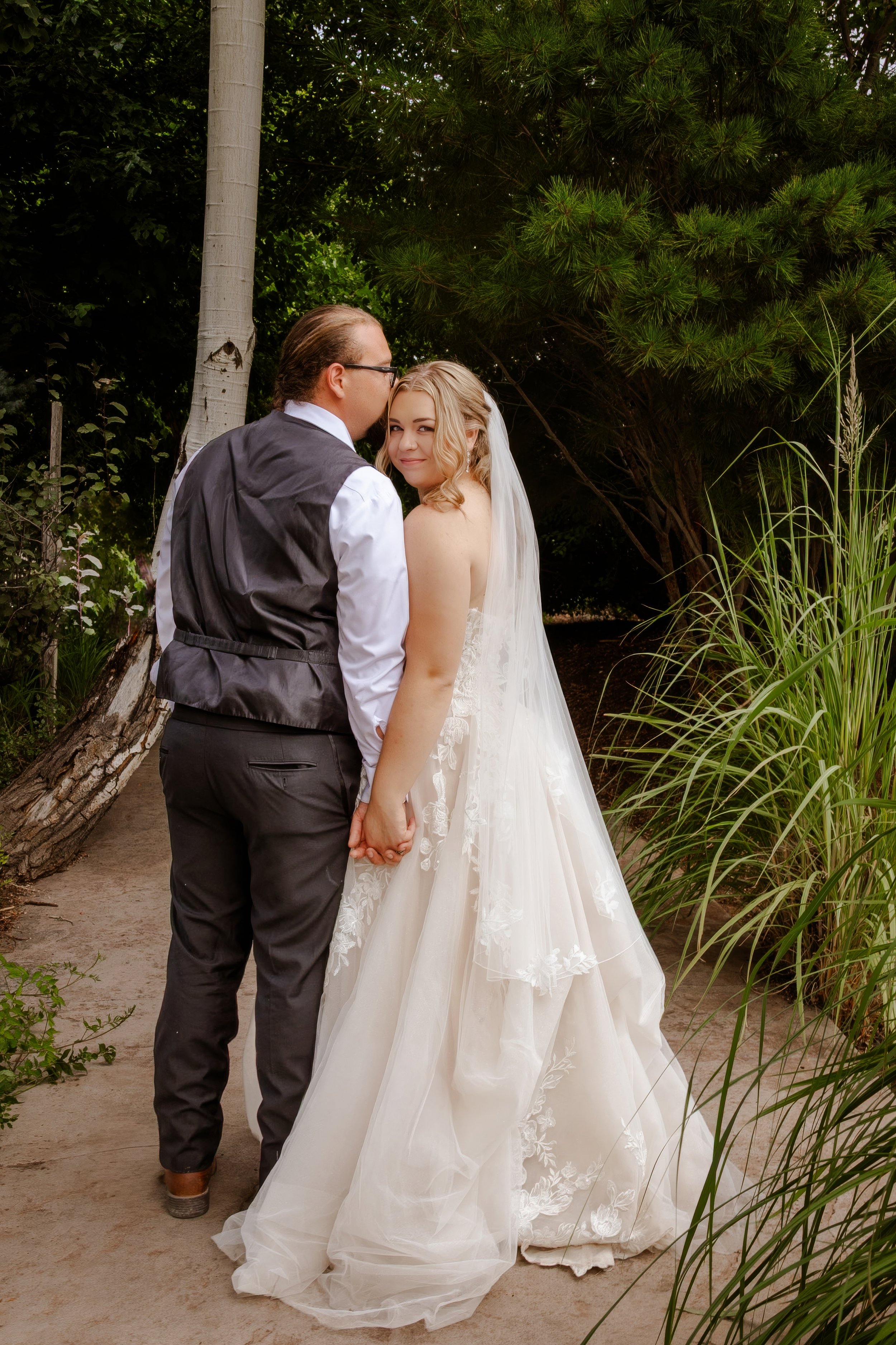 A bride and groom standing close together outdoors, holding hands, with greenery in the background. The groom is kissing the bride on the cheek, and she is smiling at the camera. The bride is wearing a white wedding gown and veil, and the groom is dr