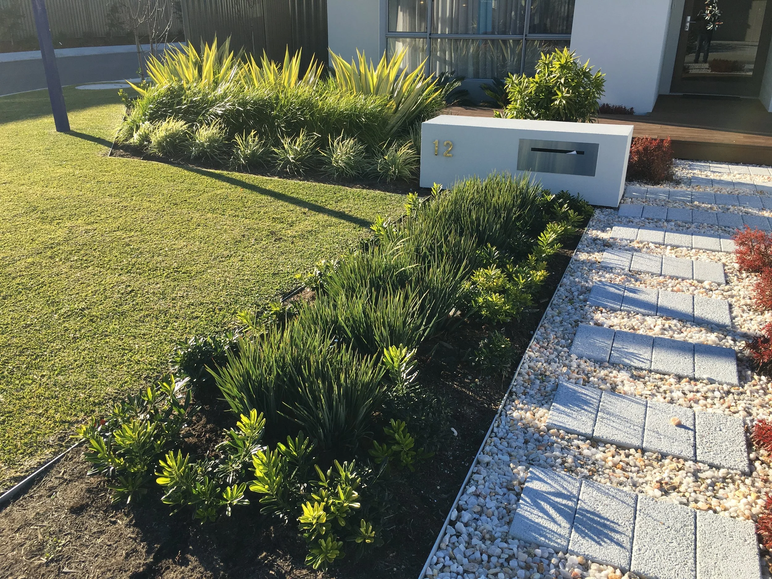 The front yard of a modern home featuring a neatly landscaped garden with lush green grass, a variety of plants, a white mailbox, and a pathway made from concrete pavers and white gravel, all cleanly defined with Hedge aluminium garden edging.