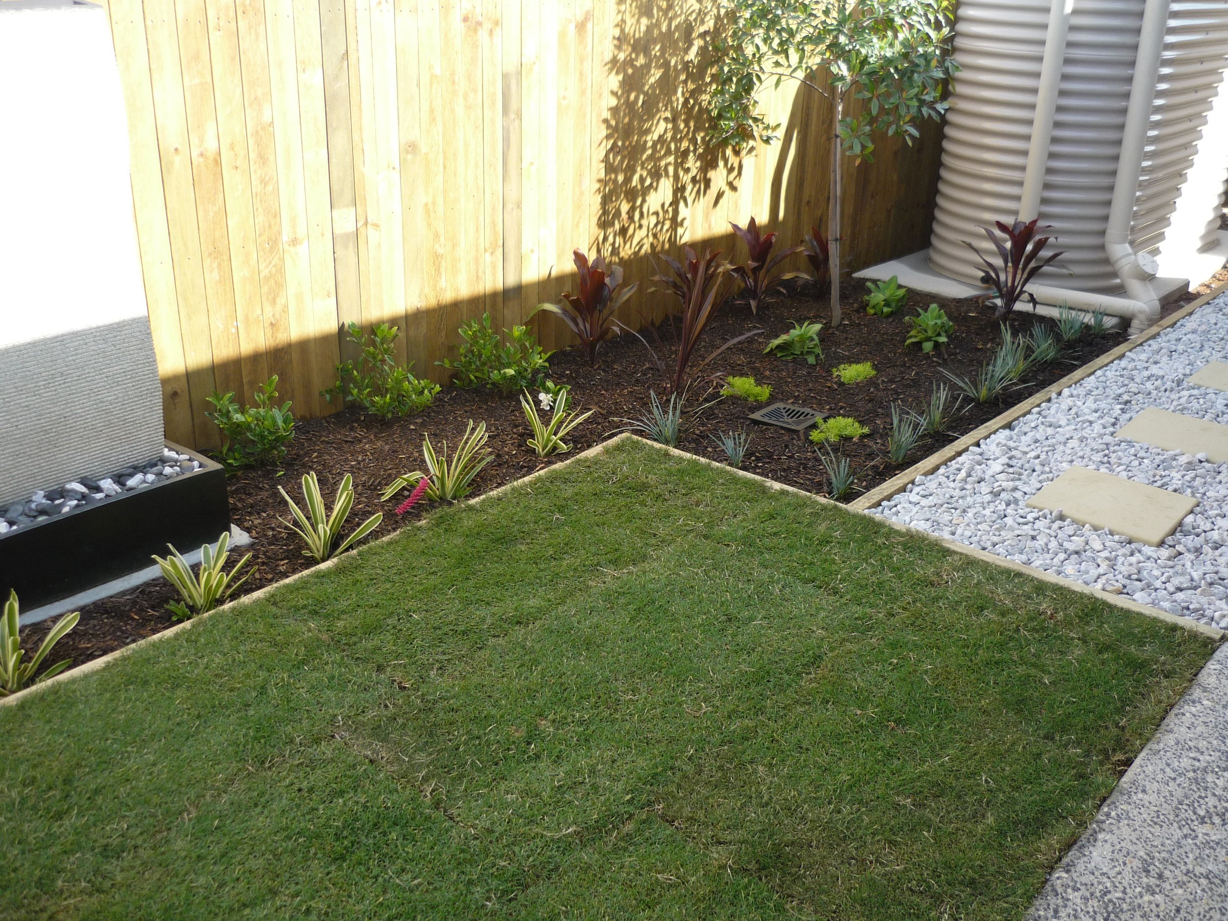A landscaped backyard with a wooden fence, ornamental plants, a grassy lawn, a rock pathway, and a drain cover.