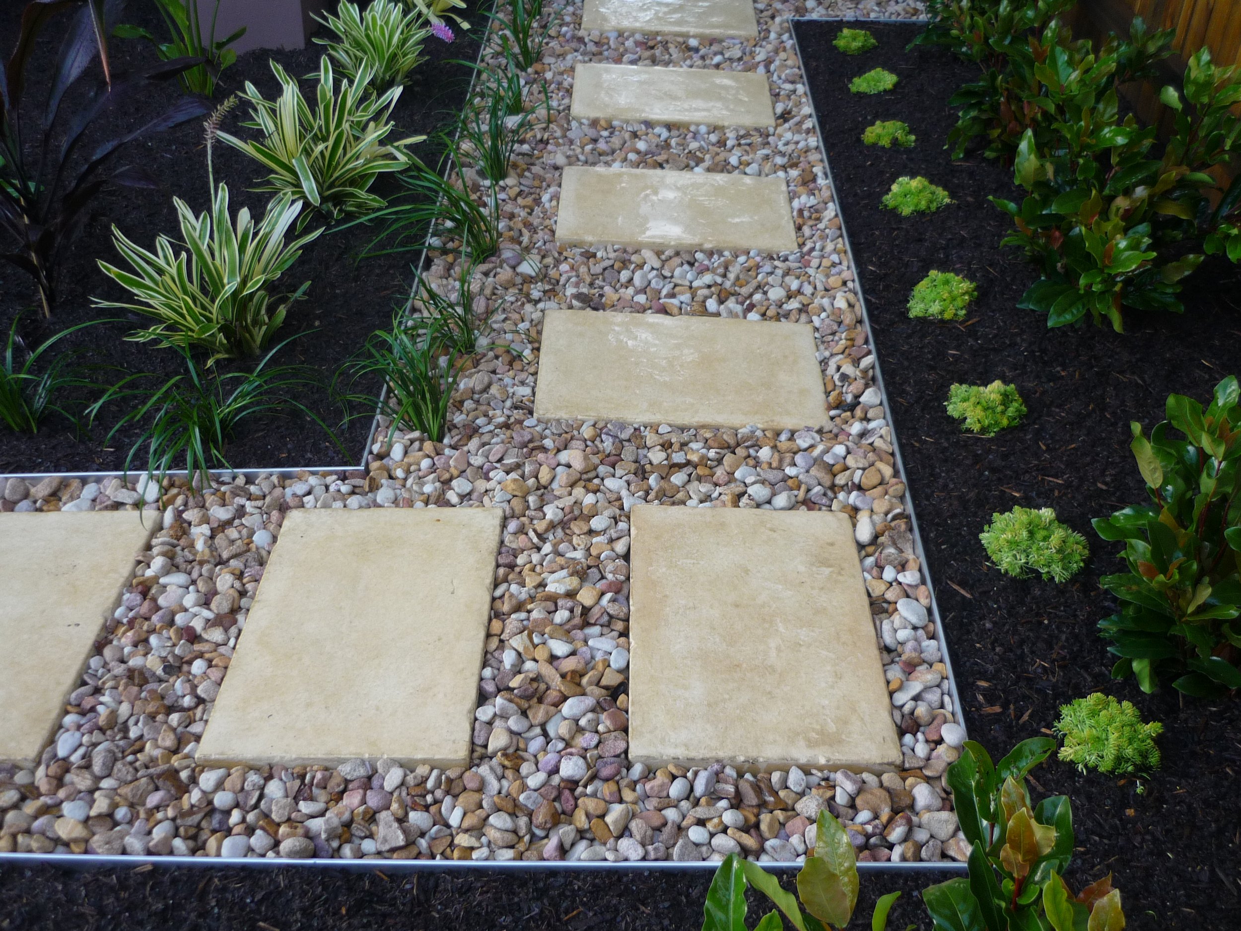 A garden pathway made of large beige stepping stones set in a bed of small multicolored pebbles, bordered by dark soil with green plants and shrubs.