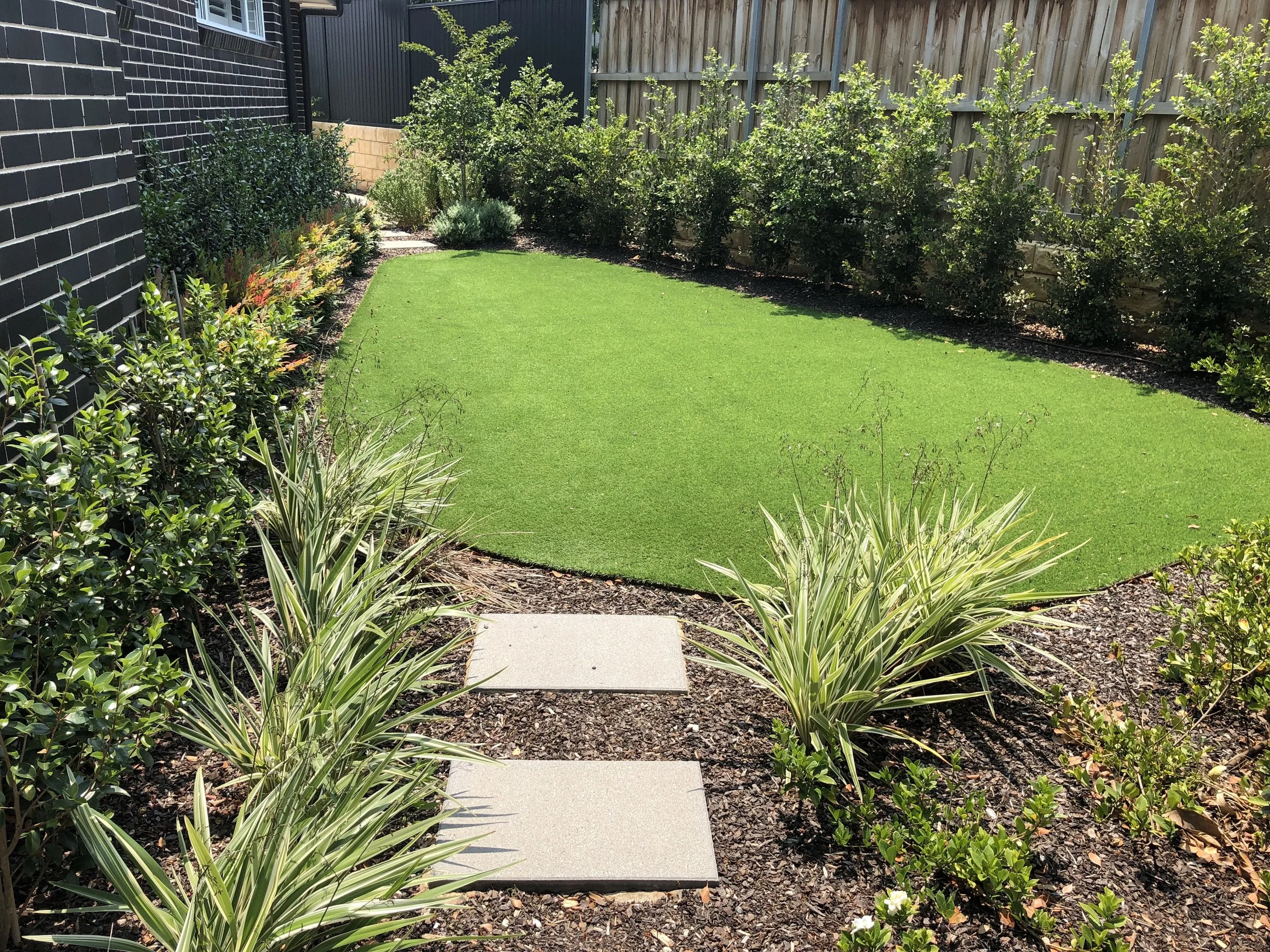 A backyard garden with a green lawn, stepping stones, bushes, and trees along a black brick wall and a wooden fence.