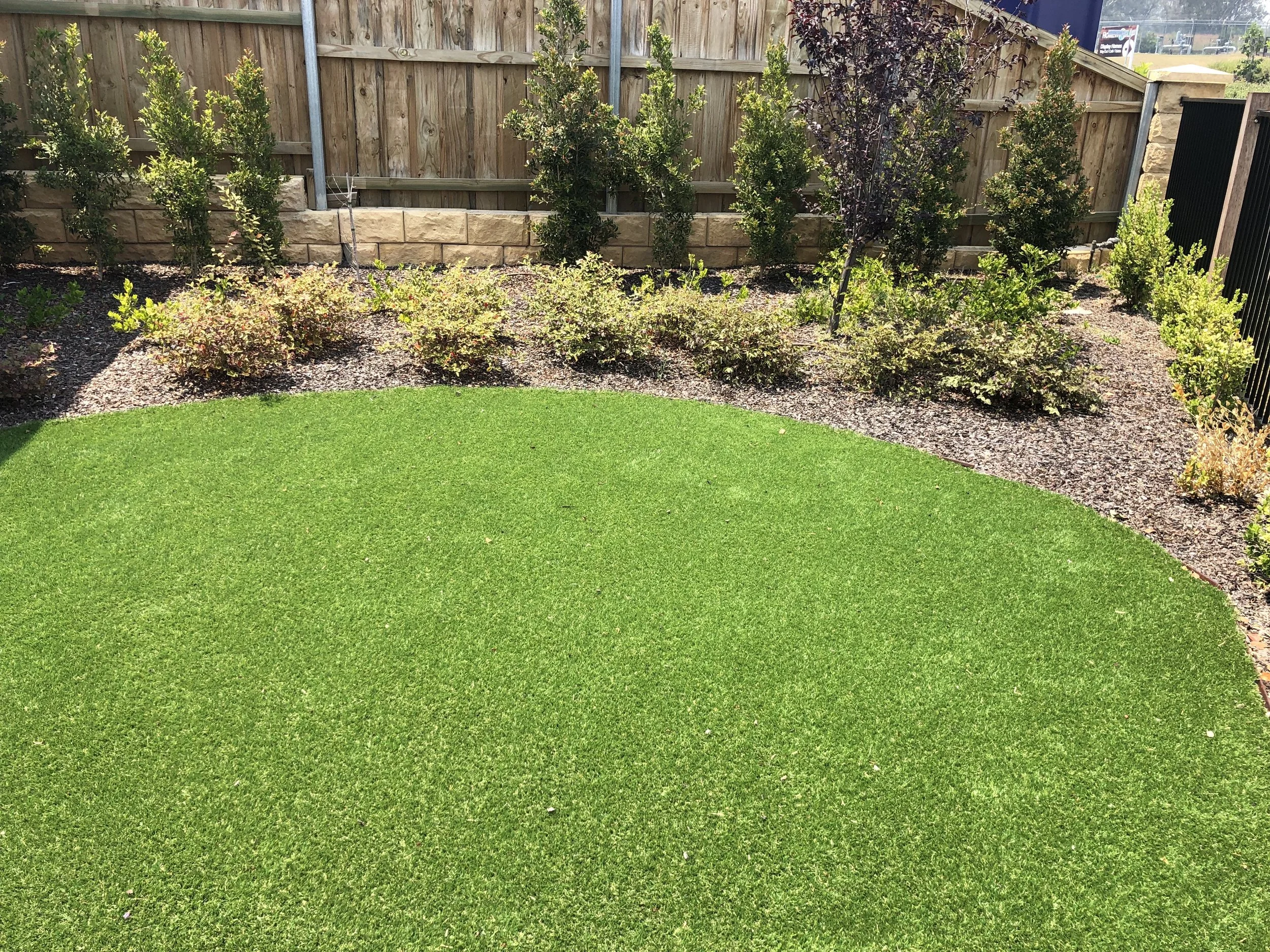 A well-maintained backyard with bright green artificial grass and a flower bed with shrubs and small trees, surrounded by a wooden privacy fence.