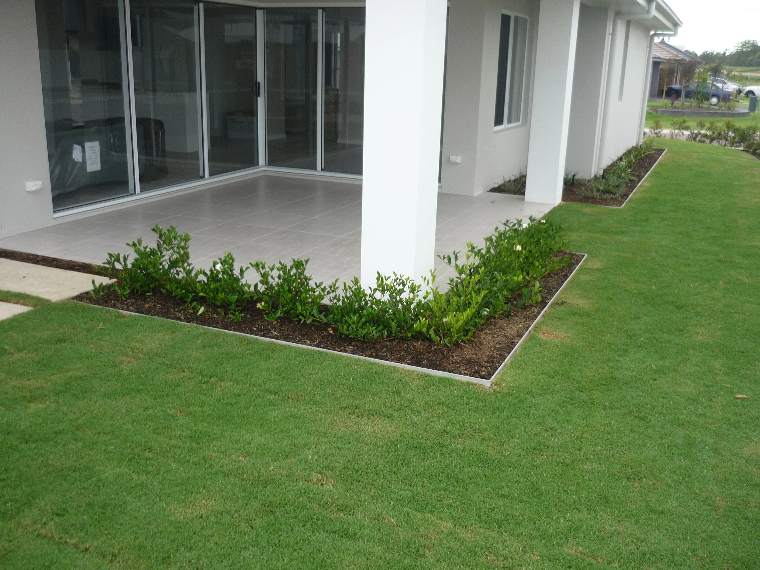 Backyard patio with sliding glass doors, a small garden bed with green shrubs, and a well-maintained grassy lawn.