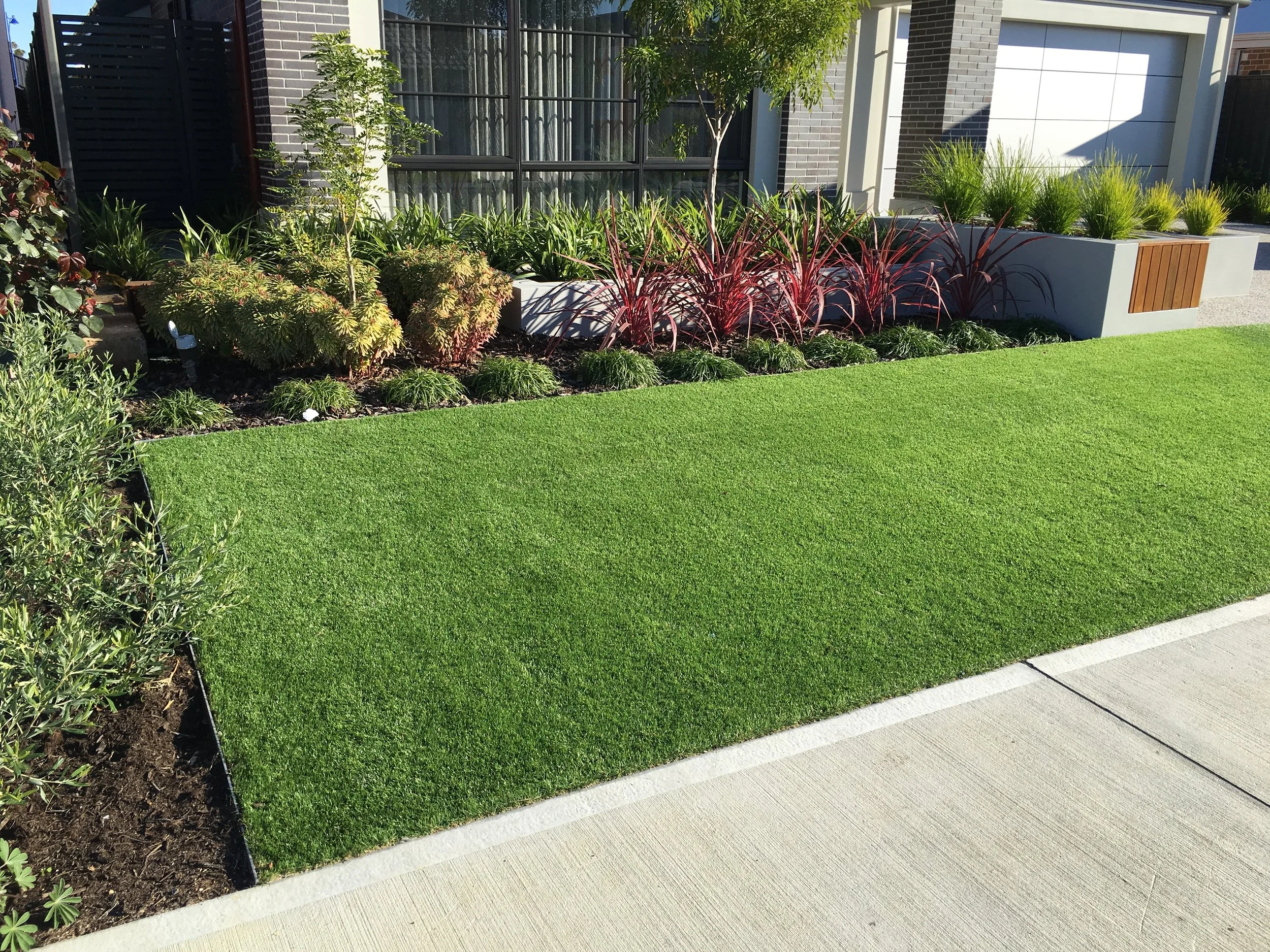 A well-manicured lawn with lush green grass beside a concrete sidewalk, bordered by a landscaped garden bed with plants and shrubs, positioned in front of a modern home with decorative planter boxes and finished with Hedge aluminium garden edging.