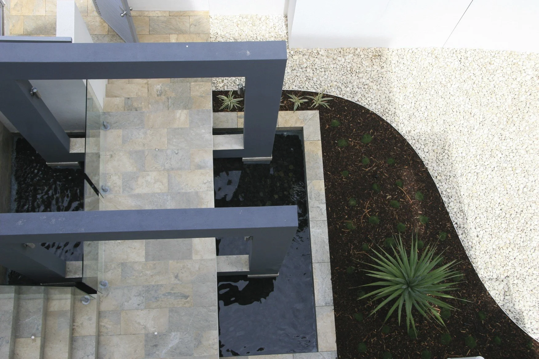 An aerial view of modern outdoor patio with stone tiles, a water feature, gray metal beams, and surrounding landscaping with small plants and white pebbles.
