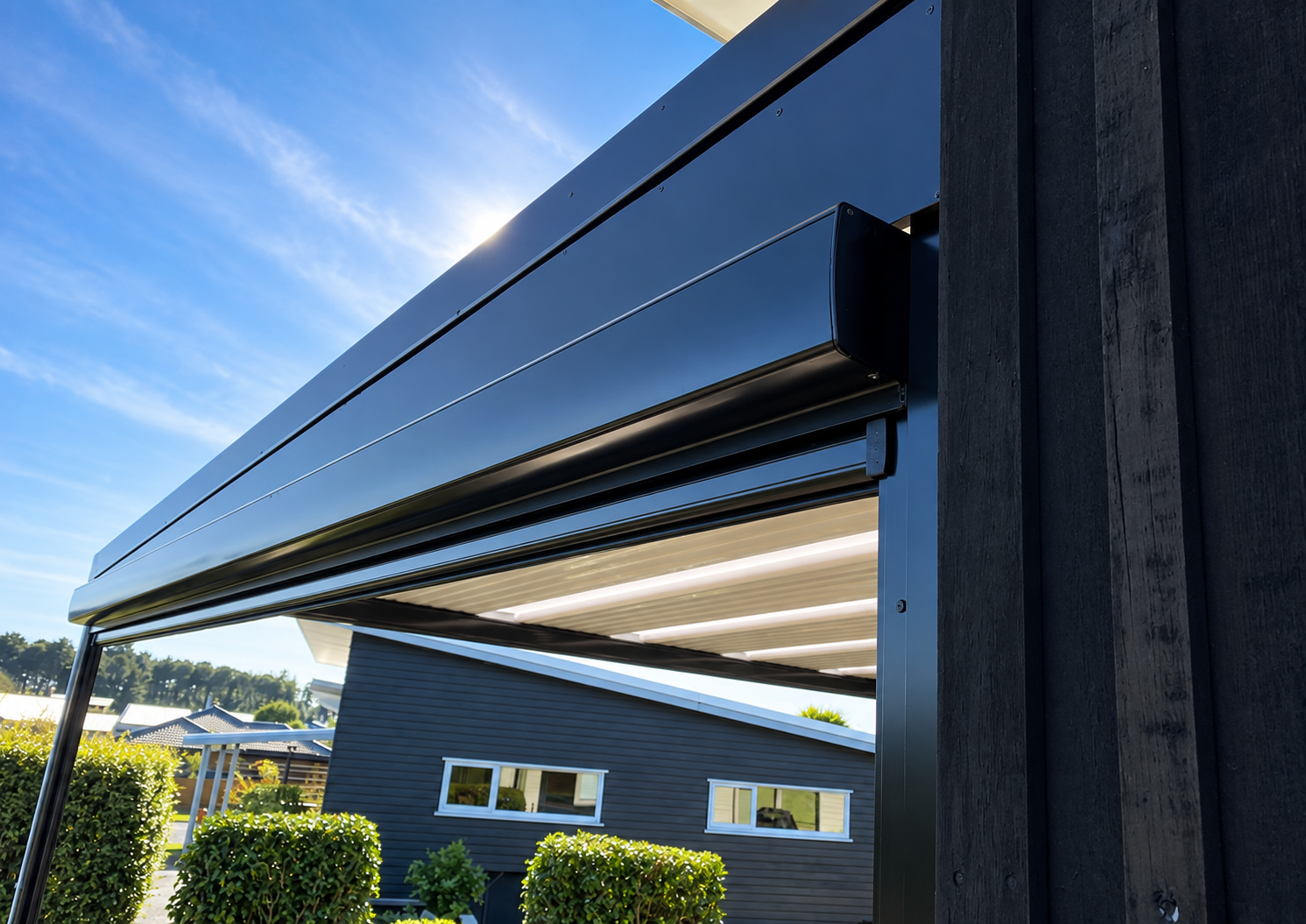 Close-up of a modern exterior structure with a black metal overhang, against a clear blue sky with the sun shining, neighboring houses, and well-maintained bushes.