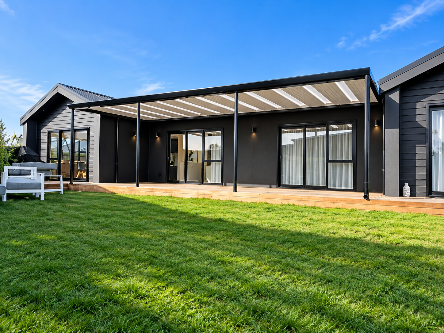 Modern black house with large sliding glass doors, a patio, and a green lawn under a blue sky.