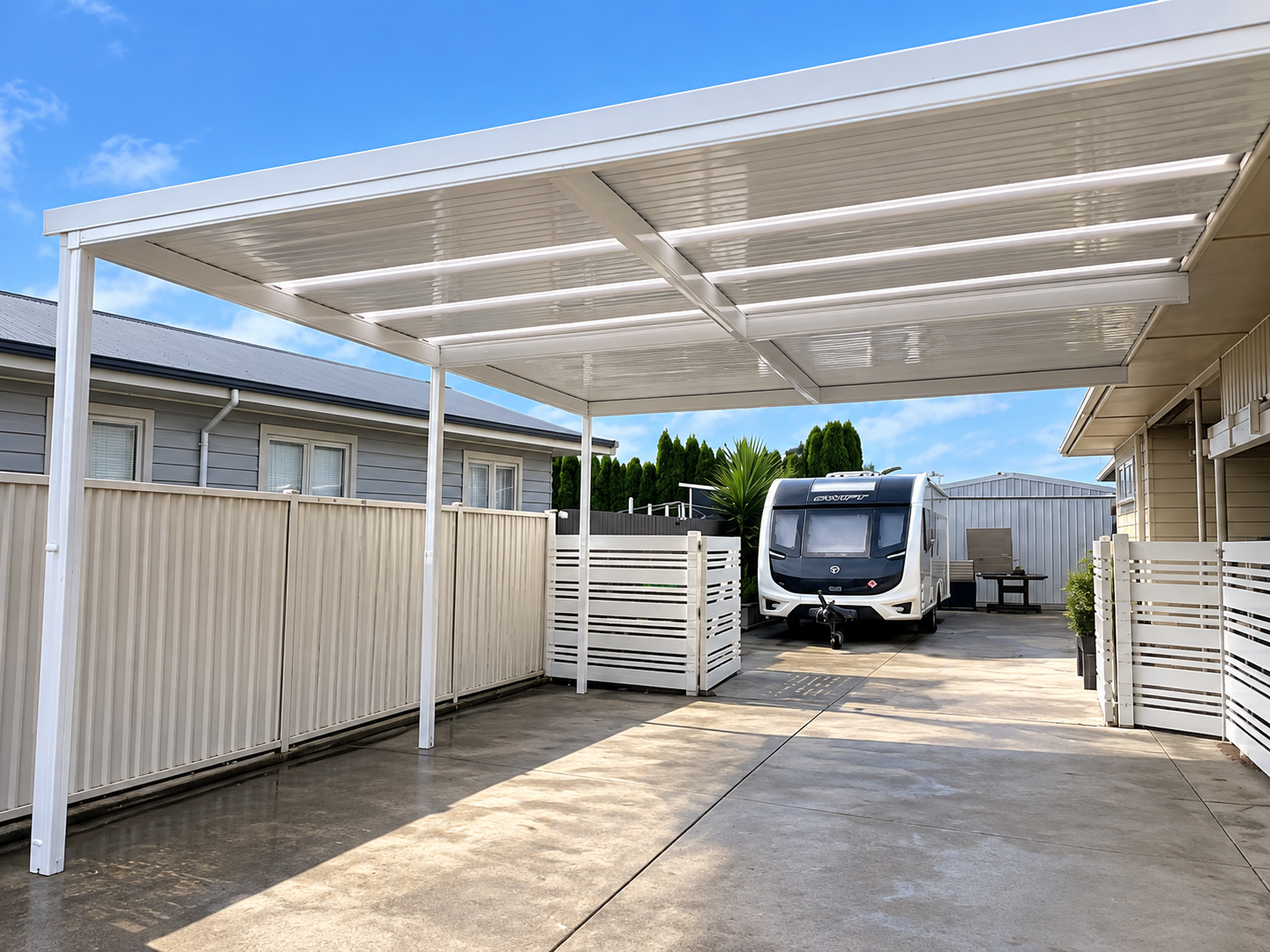 Covered carport with a recreational vehicle parked underneath, surrounded by a white fence and neighboring house on a sunny day.