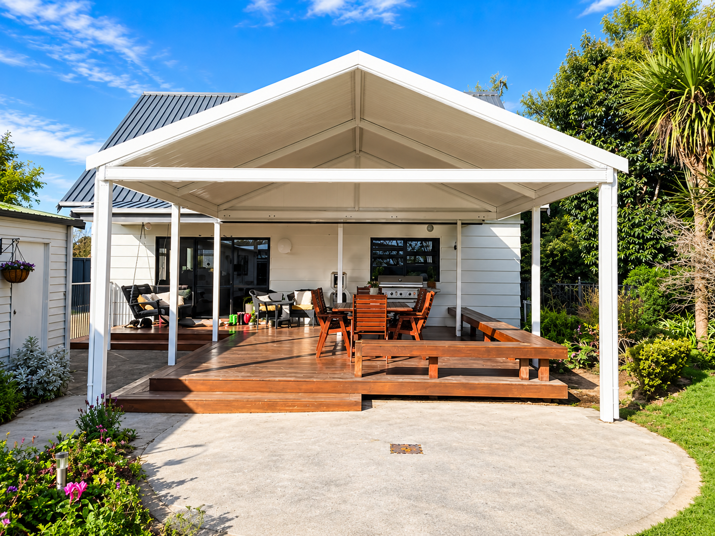 Backyard patio with wooden deck, outdoor furniture including chairs, a dining table, and a grill, surrounded by greenery and trees under a blue sky.