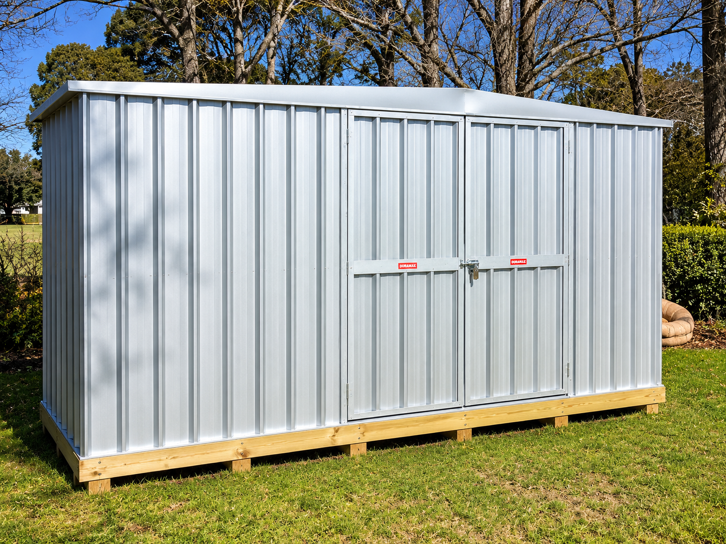 A silver metal shed with double doors, set on wooden supports in a grassy yard with trees and bushes in the background under a clear blue sky.