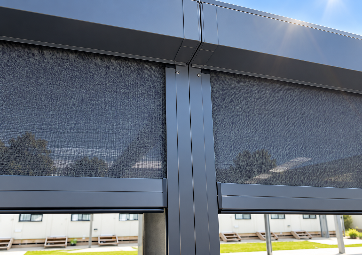 Close-up of a modern building with dark gray metal panels and mesh screens, with a clear blue sky and some trees reflected in the surfaces.