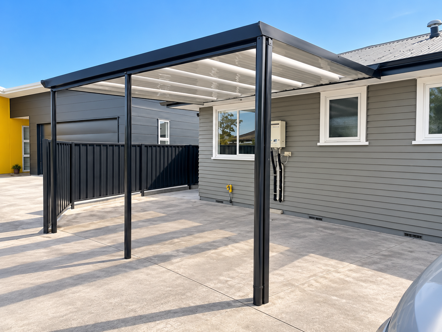 Empty outdoor patio area with a metal-framed pergola attached to the side of a gray house, with concrete flooring and neighboring modern houses in the background.