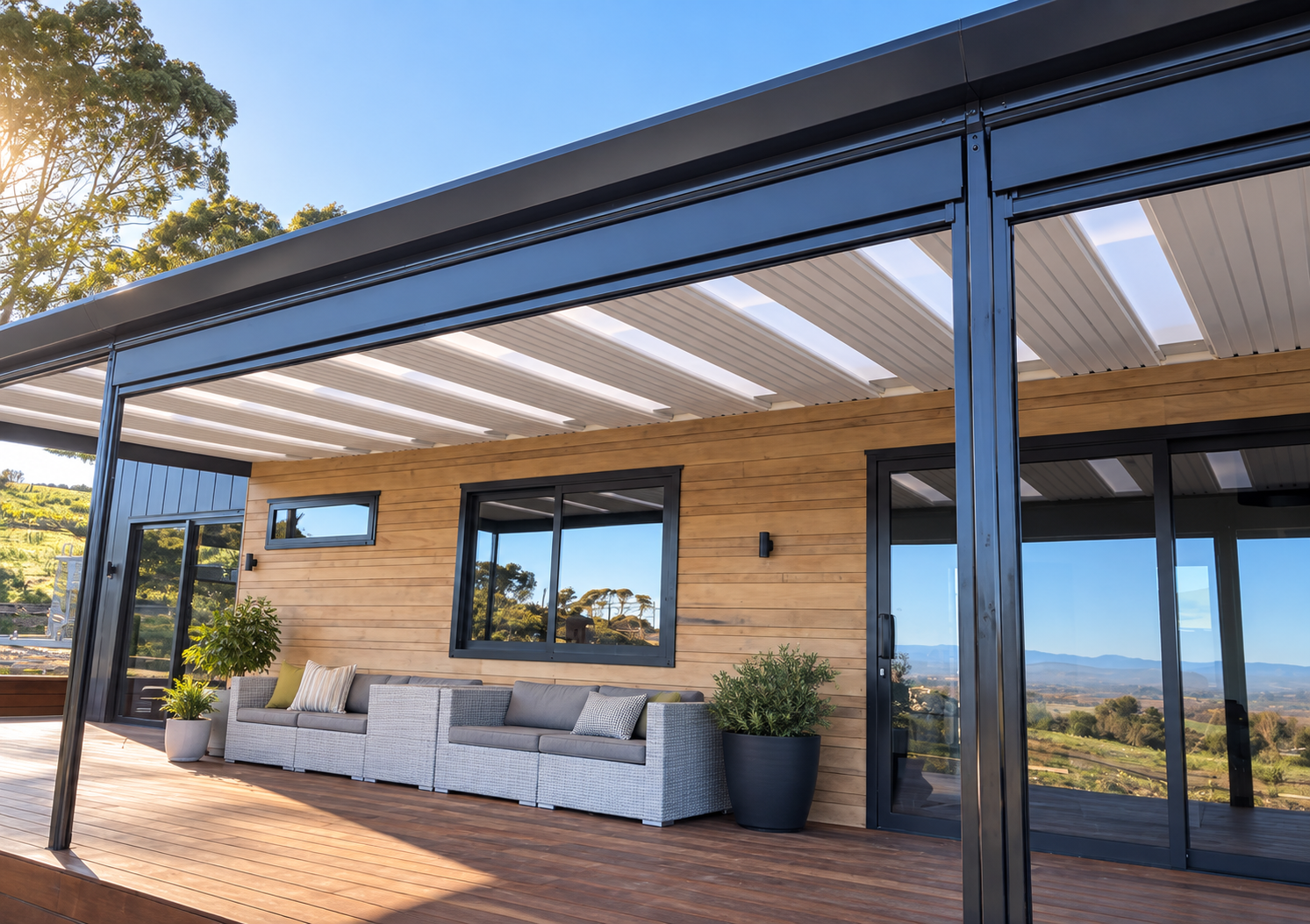 Modern house with wooden exterior and glass sliding doors, outdoor sitting area with sofas, potted plants, and rolling hills in the background.