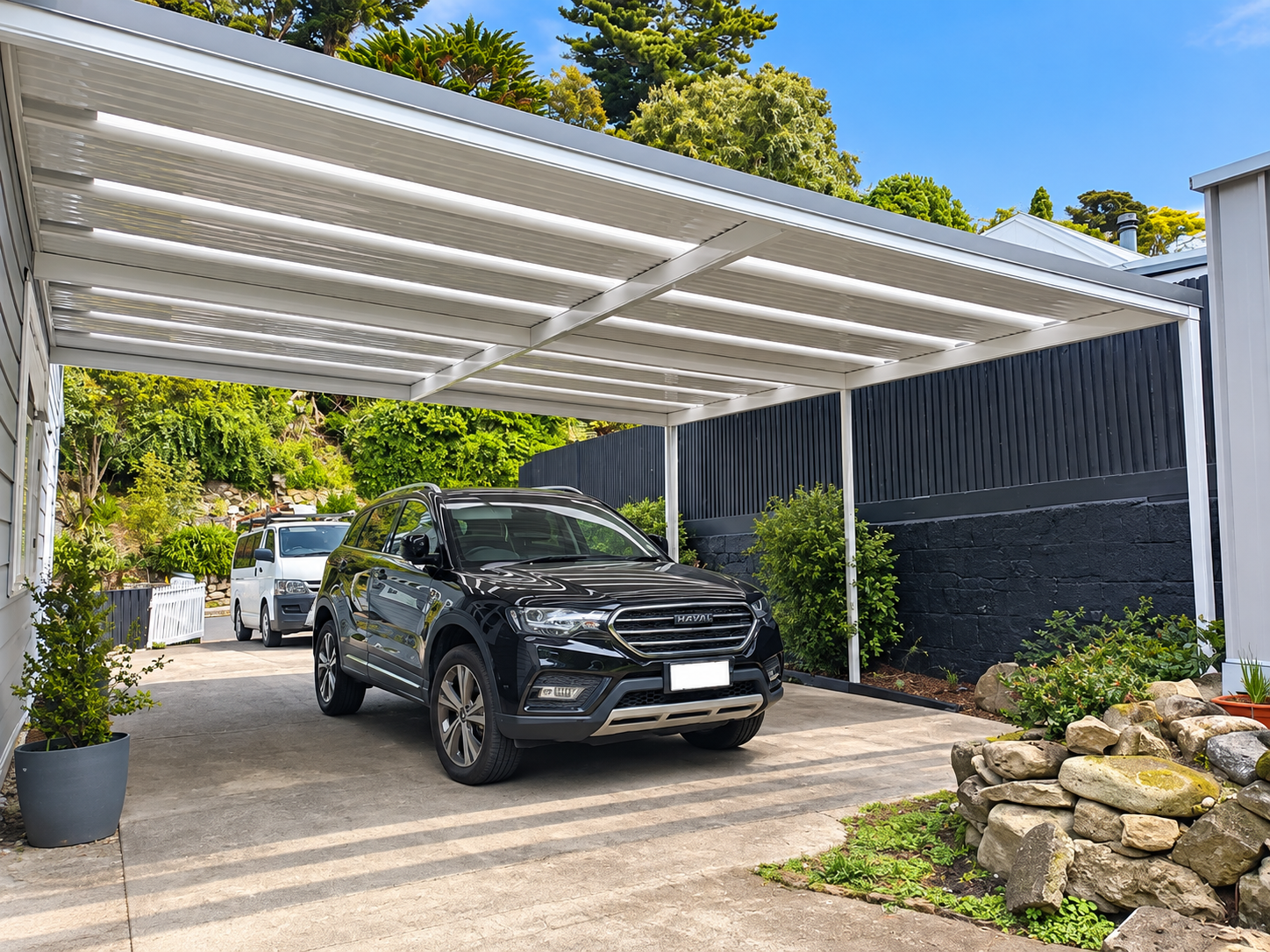 A black SUV and a white vehicle parked under a white metal carport with horizontal slats, in a residential driveway. There are green shrubs and trees in the background, along with a black fence on one side and a stone rock garden on the other.