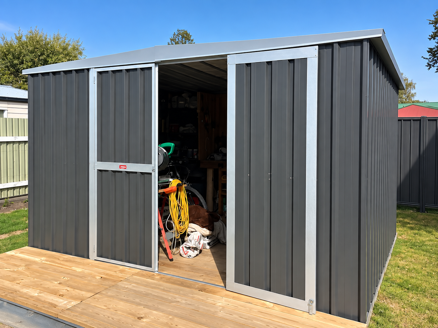 Black metal storage shed with open sliding door, revealing gardening tools, a ladder, and various equipment inside, on a wooden deck with grass and fences in the background.