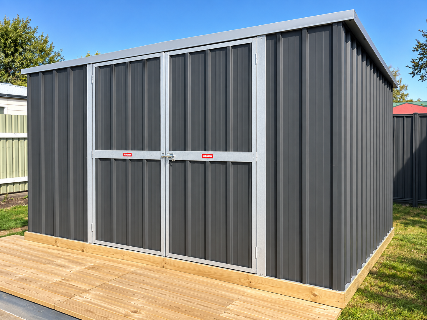 A dark gray metal storage shed with double doors secured by a latch, set on a wooden platform in a backyard with a green lawn and a white fence, under a blue sky.