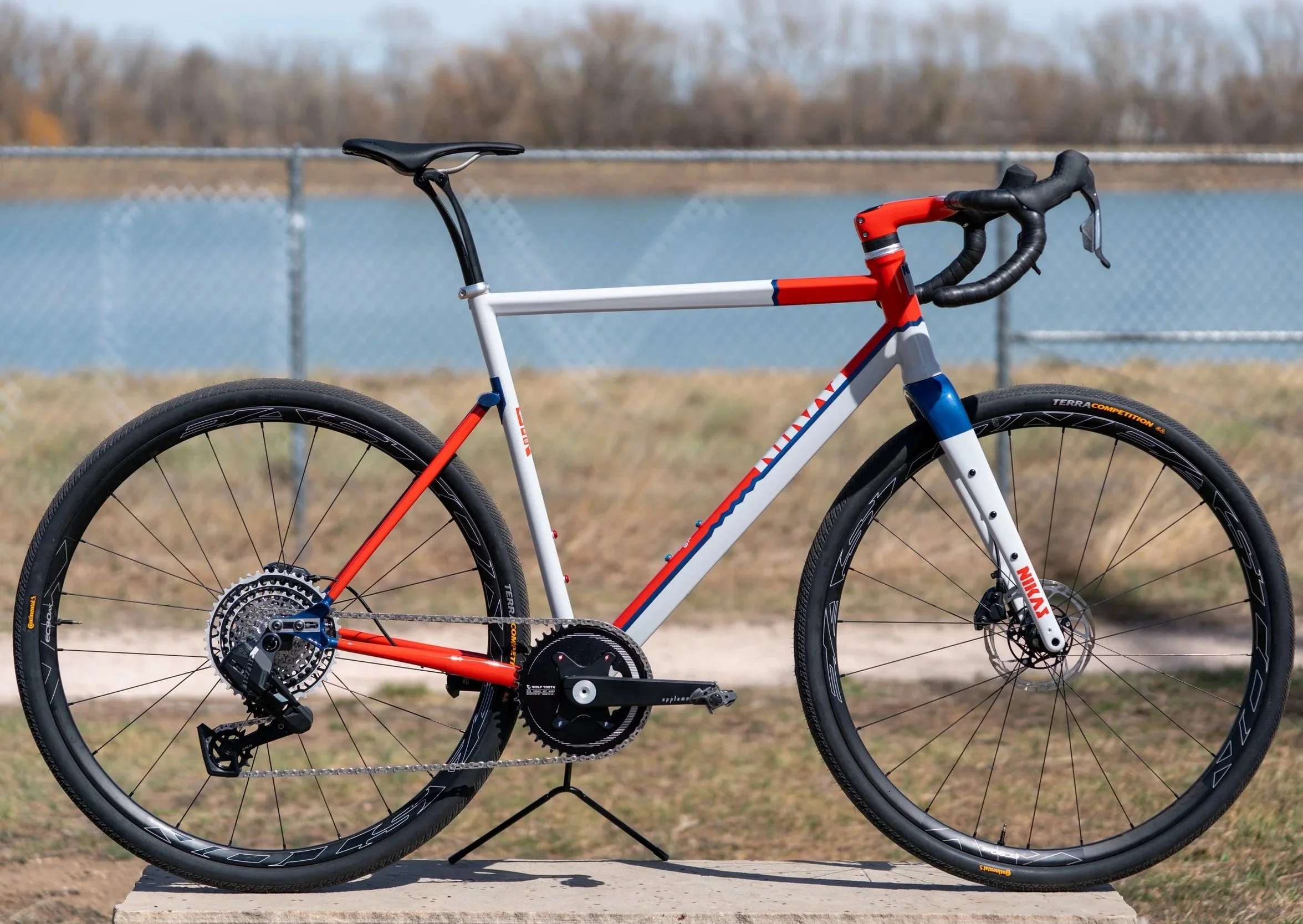 A modern racing bicycle with a white, red, and blue frame, black tires, and a black saddle, positioned outdoors on a concrete block with a fence and water body in the background.